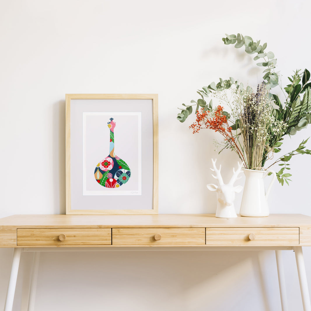 A framed fine art print, by Lis Gonçalves. Open edition, signed by the artist (printed in Hahnemühle matt fibre paper) featuring a colorful Portuguese guitar, leaning on a light wood console table next to a white ceramic deer figurine and a vase of green foliage, set against a plain white wall.