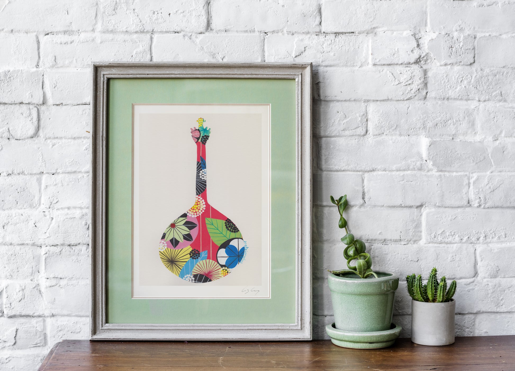 A framed fine art print, by Lis Gonçalves. Open edition, signed by the artist (printed in Hahnemühle matt fibre paper) featuring a colorful Portuguese guitar on a wooden surface with potted plants against a white brick wall.