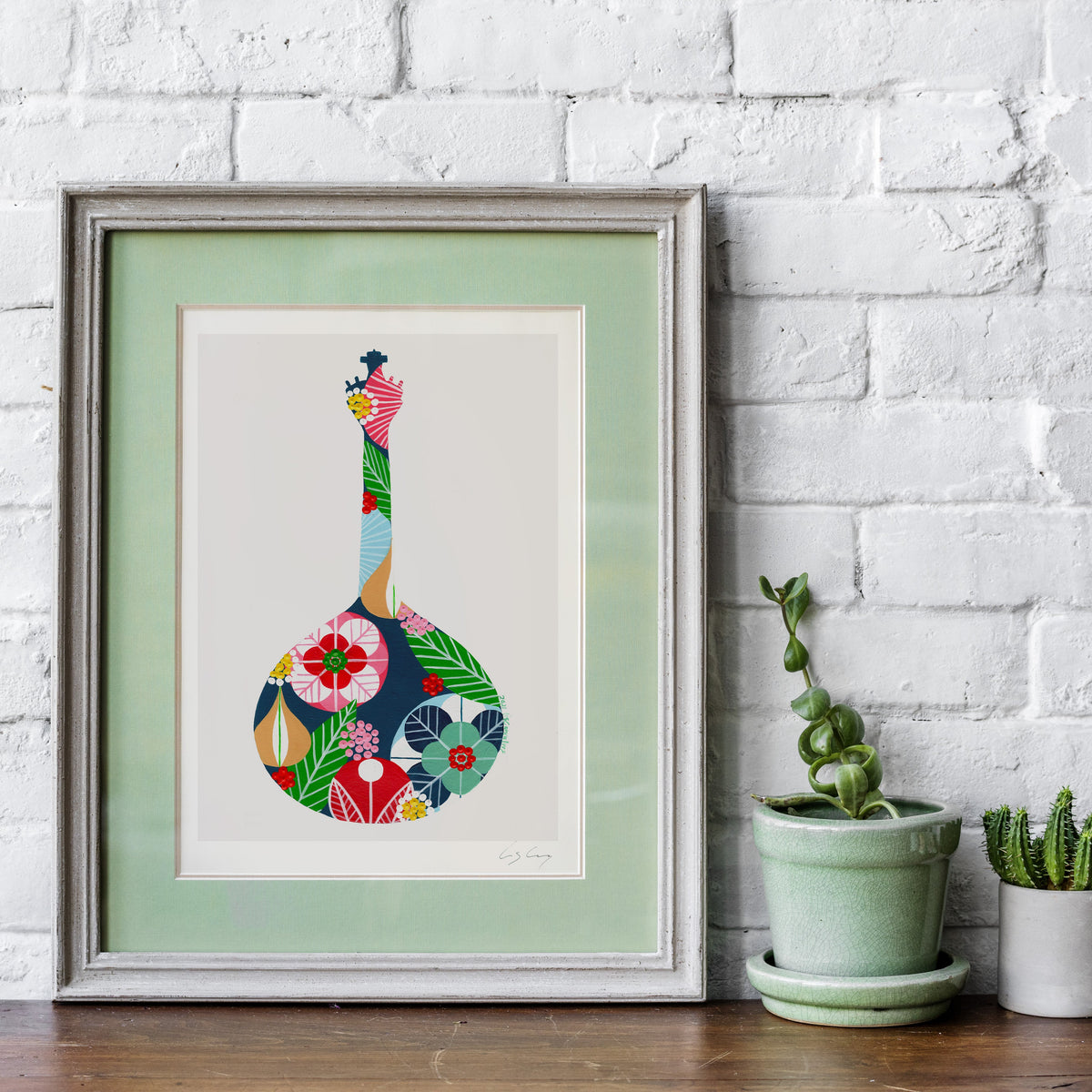 A framed fine art print, by Lis Gonçalves. Open edition, signed by the artist (printed in Hahnemühle matt fibre paper) featuring a colorful Portuguese guitar, displayed on a wooden surface against a white brick wall next to two potted plants.