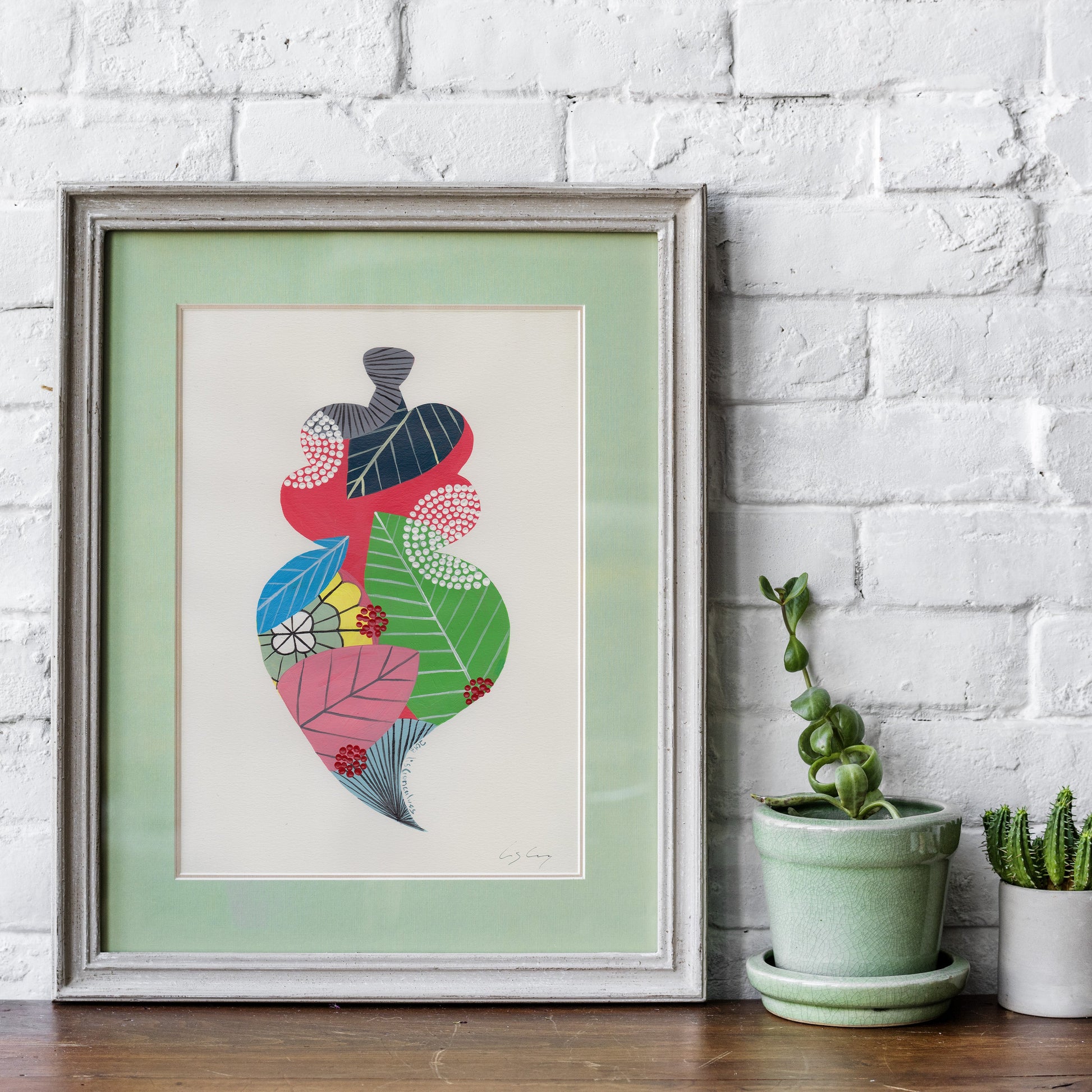 A framed fine art print, by Lis Gonçalves. Open edition, signed by the artist (printed in Hahnemühle matt fibre paper) featuring a colorful Portuguese Viana heart, leaning against a white brick wall on a wooden surface next to two small potted succulents.