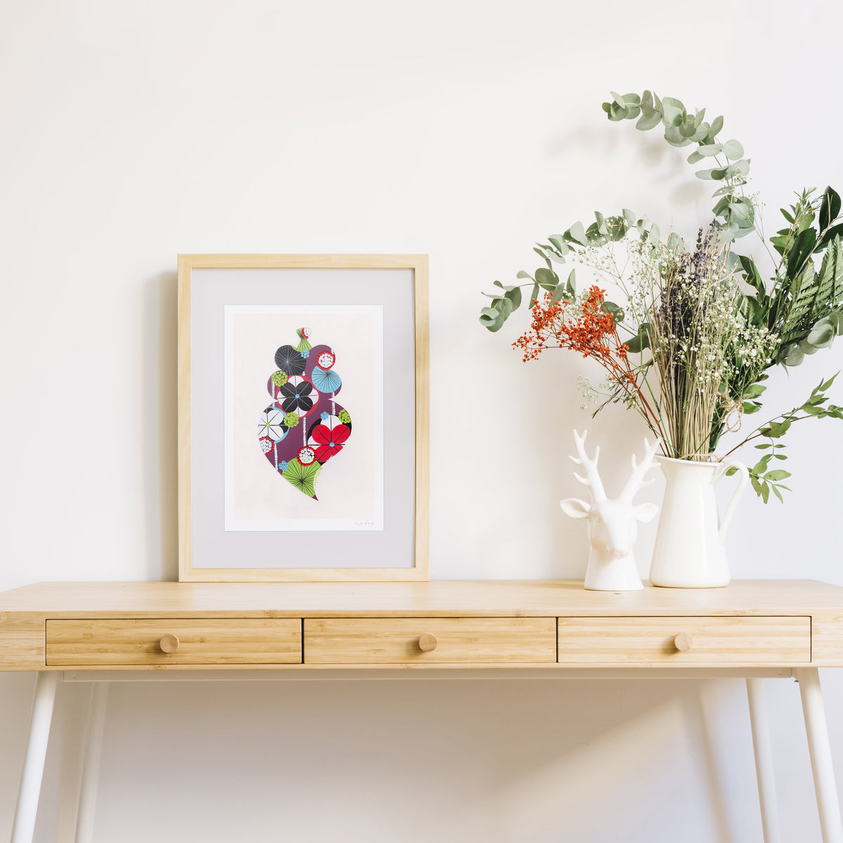 A framed fine art print, by Lis Gonçalves. Open edition, signed by the artist (printed in Hahnemühle matt fibre paper) featuring a colorful Portuguese Viana heart, styled on a light wood console table next to a white ceramic deer figurine and a vase of green and orange dried flowers against a white wall.