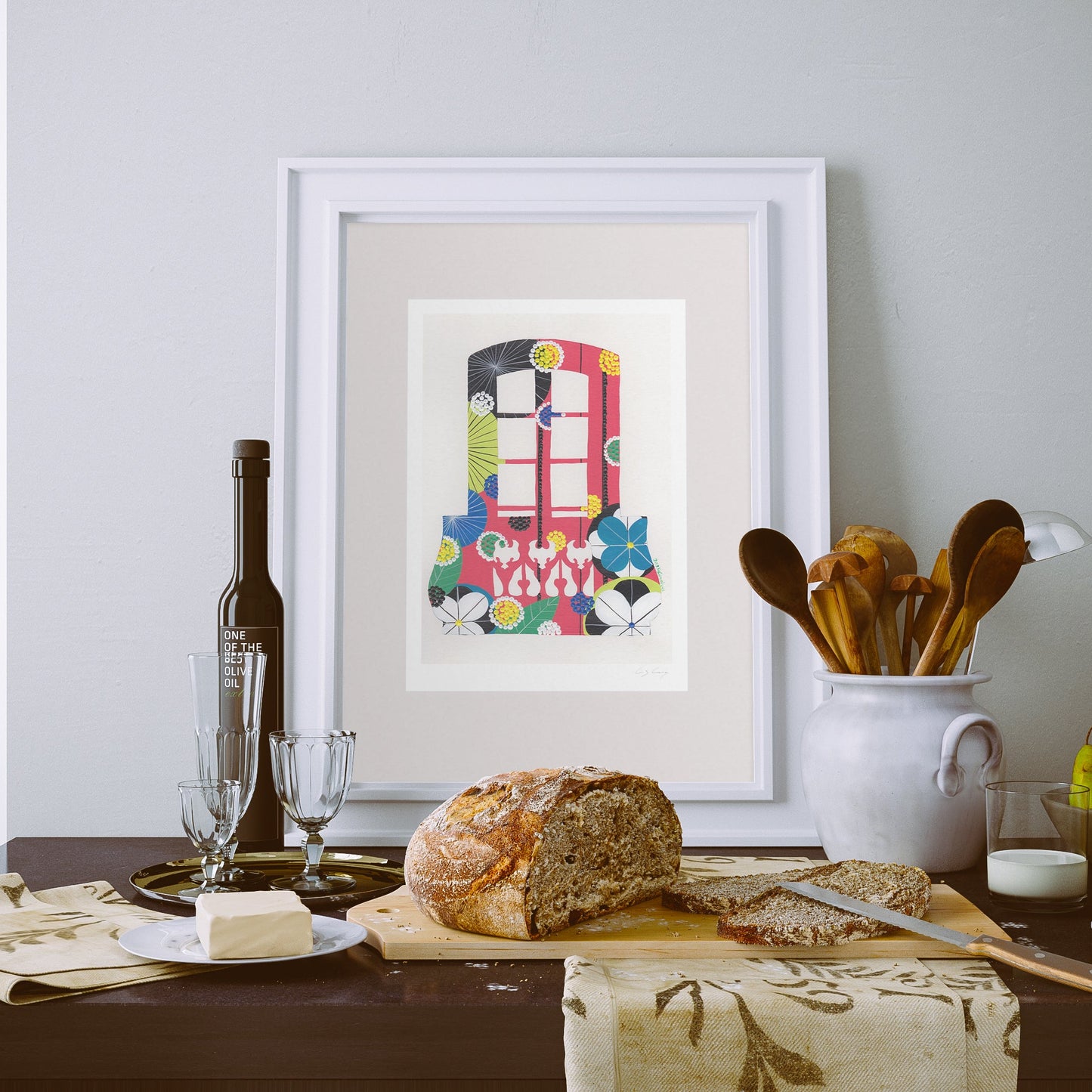 A framed fine art print, by Lis Gonçalves. Open edition, signed by the artist. Printed in Hahnemühle matt fibre paper; featuring a colorful illustration of a pink Lisbon balcony with flowers, displayed on a dining table alongside a loaf of bread, butter, wine, and a ceramic container of wooden utensils.
