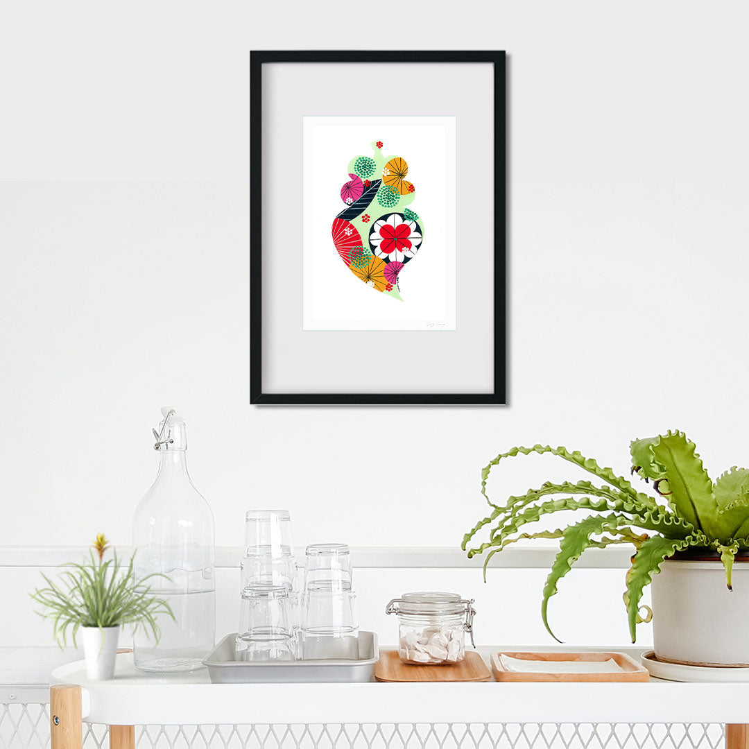 A framed fine art print, by Lis Gonçalves. Open edition, signed by the artist (printed in Hahnemühle matt fibre paper) featuring a colorful Portuguese Viana heart on a white wall with plants and decor below.