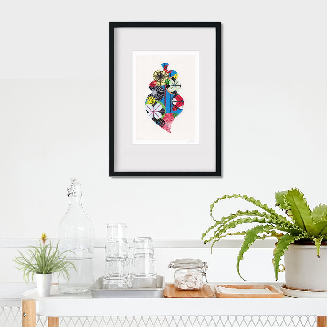 A framed fine art print, by Lis Gonçalves. Open edition, signed by the artist (printed in Hahnemühle matt fibre paper) featuring a colorful Portuguese Viana heart on a white wall above a kitchen counter with plants and glasses.