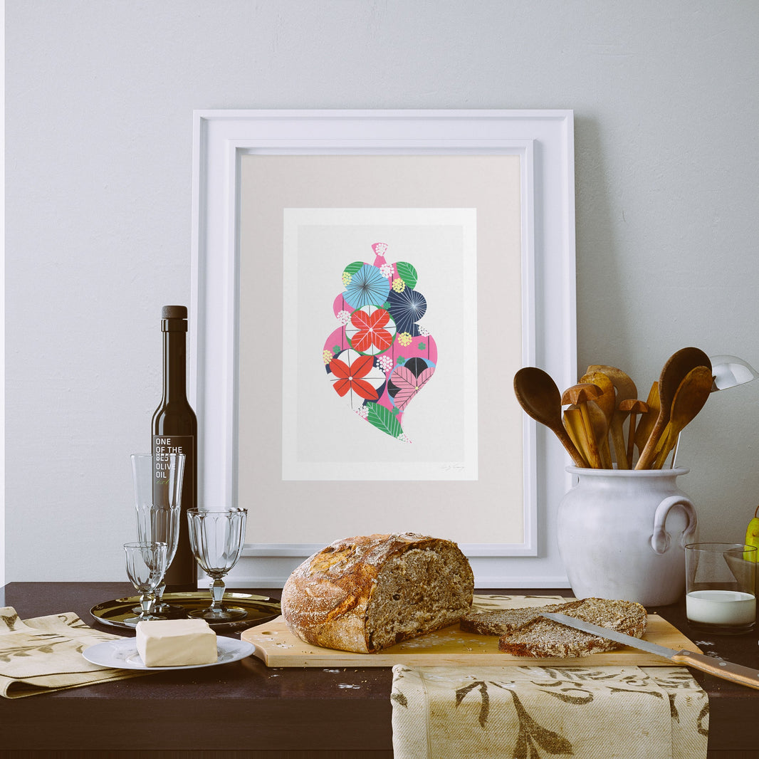 A framed fine art print, by Lis Gonçalves. Open edition, signed by the artist (printed in Hahnemühle matt fibre paper) featuring a colorful Portuguese Viana heart, displayed on a table with a loaf of bread, butter, wine glasses, and wooden utensils against a light gray wall.