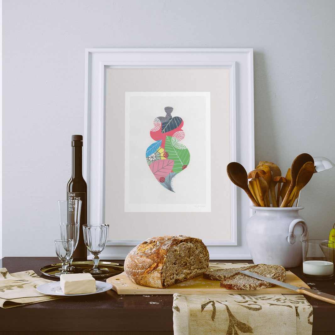 A framed fine art print, by Lis Gonçalves. Open edition, signed by the artist (printed in Hahnemühle matt fibre paper) featuring a colorful Portuguese Viana heart  on a table setting with bread, wine, and utensils against a wall.