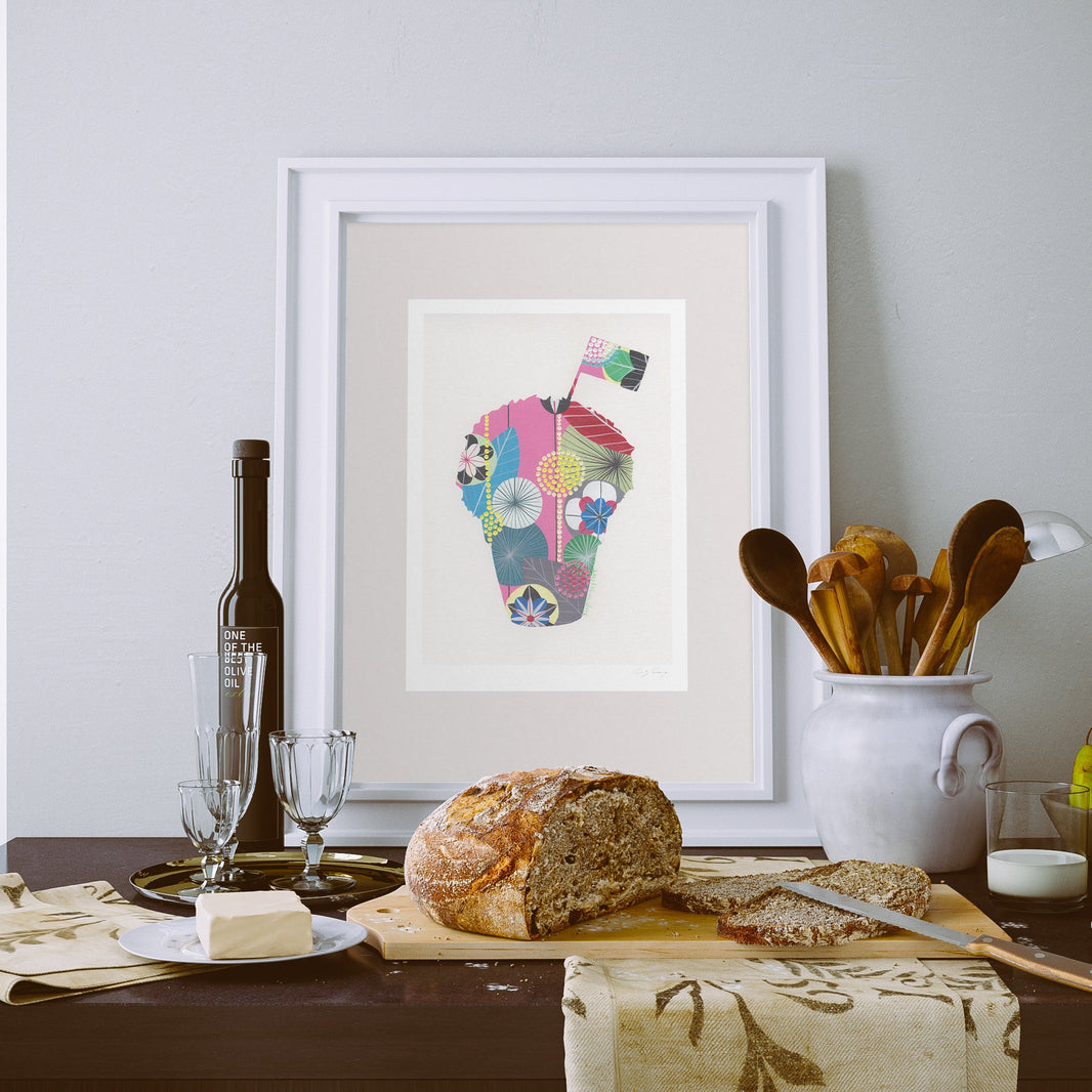 A framed fine art print, by Lis Gonçalves. Open edition, signed by the artist (printed in Hahnemühle matt fibre paper) featuring a colorful, patterned manjerico illustration leaning against a light gray wall on a dining table. The table is styled with a loaf of bread, butter, wine glasses, a bottle, and a white ceramic pot holding wooden utensils.