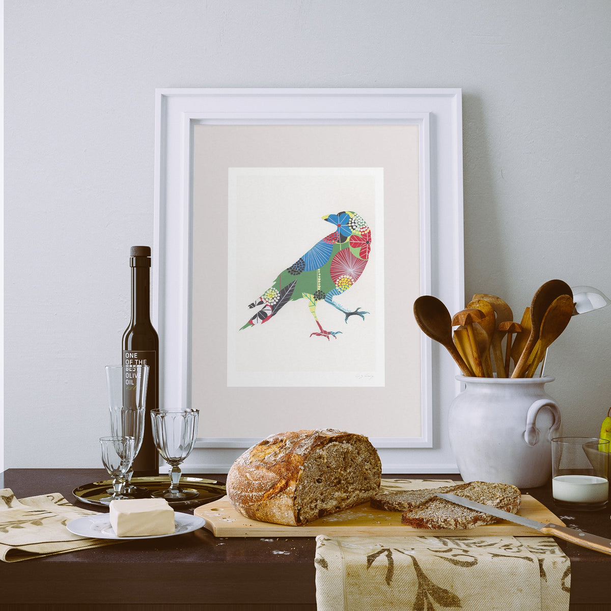 A framed fine art print, by Lis Gonçalves. Open edition, signed by the artist (printed in Hahnemühle matt fibre paper) featuring a colorful raven on a white background, styled on a dining table with a loaf of bread, wine glasses, a bottle, and a white ceramic container of wooden utensils.