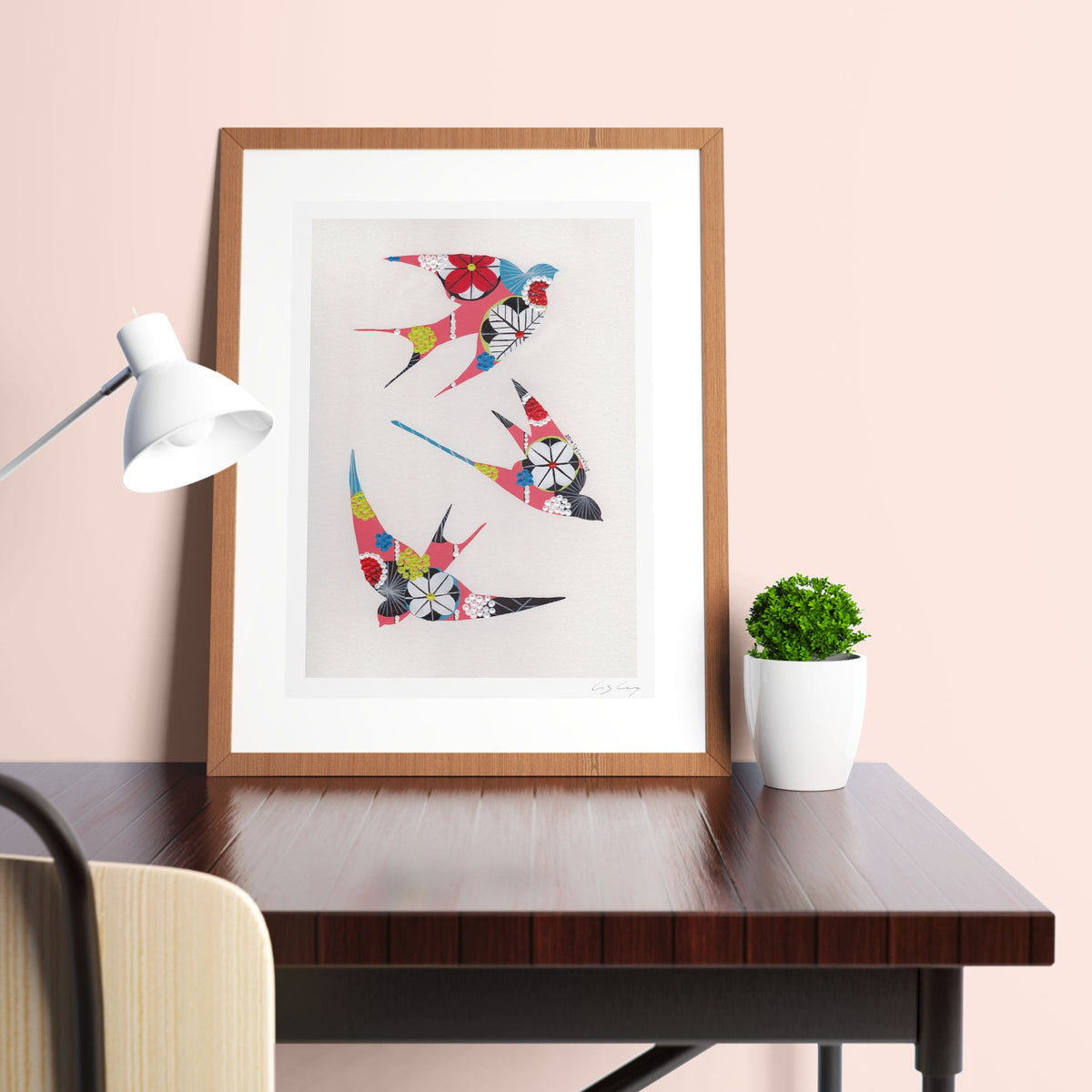 A framed fine art print, by Lis Gonçalves. Open edition, signed by the artist (printed in Hahnemühle matt fibre paper) featuring colorful swallows on a desk with a lamp and plant against a pink wall