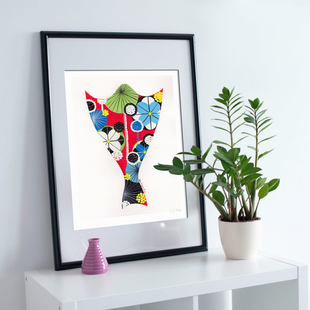 A framed fine art print, by Lis Gonçalves. Open edition, signed by the artist (printed in Hahnemühle matt fibre paper) featuring a colorful bacalhau (cod fish) in a black frame, styled on a white shelf next to a potted green plant and a small pink vase against a light gray wall.