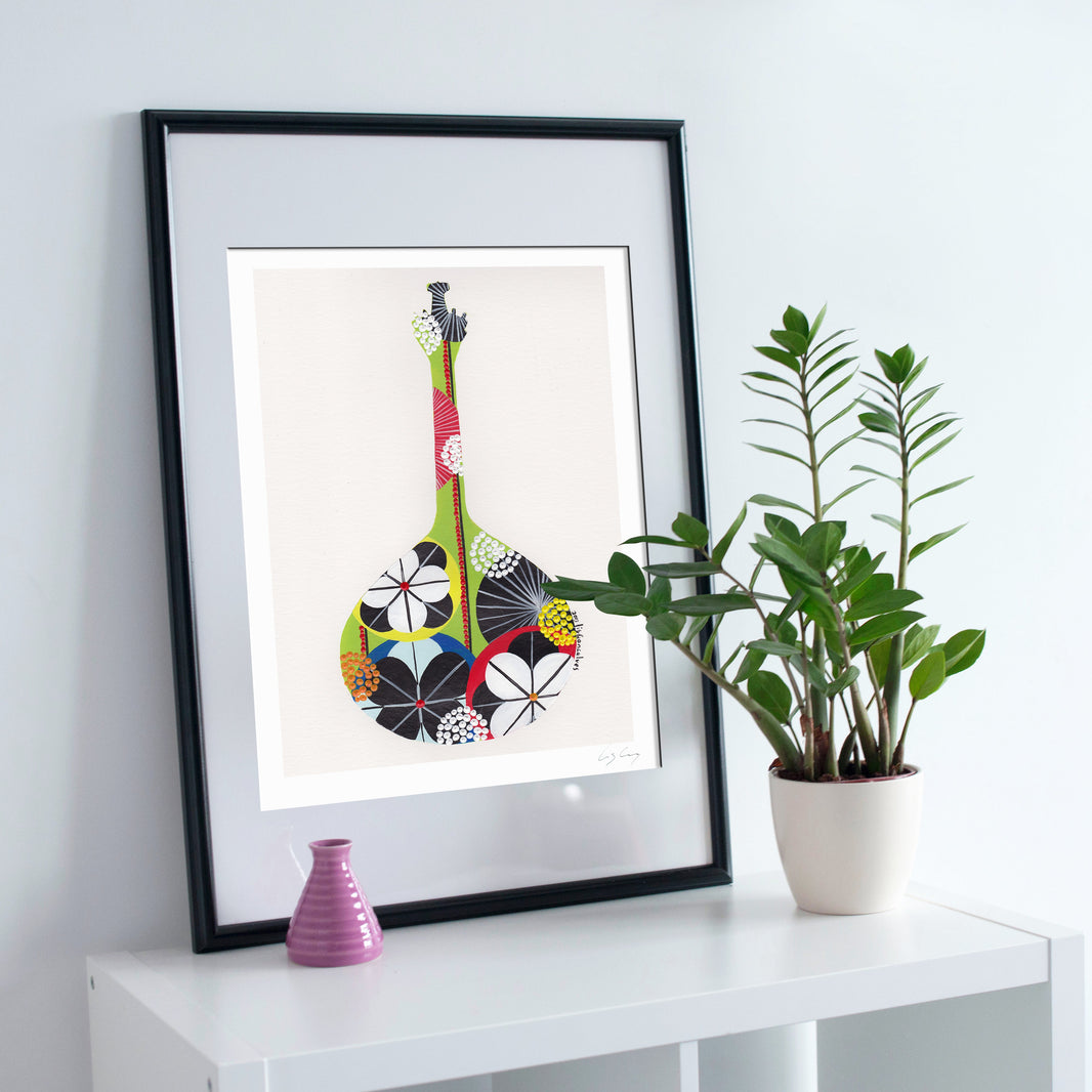 A framed fine art print, by Lis Gonçalves. Open edition, signed by the artist (printed in Hahnemühle matt fibre paper) featuring a colorful Portuguese guitar, styled on a white shelf next to a potted green plant and a small pink vase against a light gray wall.