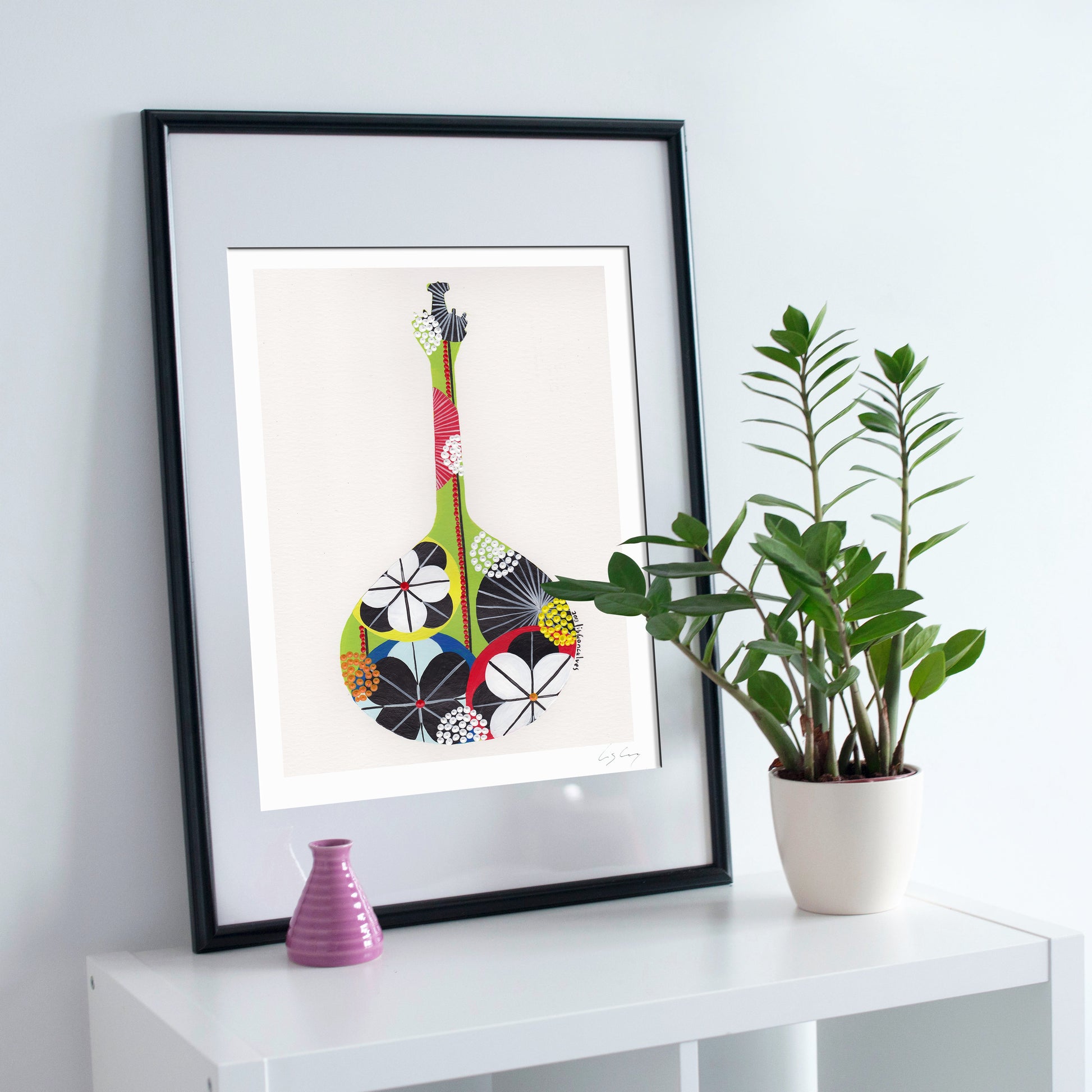 A framed fine art print, by Lis Gonçalves. Open edition, signed by the artist (printed in Hahnemühle matt fibre paper) featuring a colorful Portuguese guitar, styled on a white shelf next to a potted green plant and a small pink vase against a light gray wall.