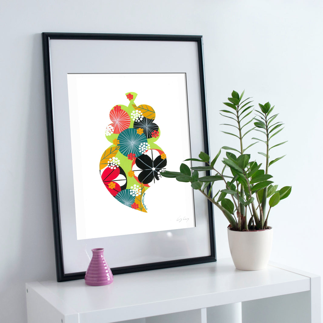 A framed fine art print, by Lis Gonçalves. Open edition, signed by the artist (printed in Hahnemühle matt fibre paper) featuring a colorful Portuguese Viana heart on a white wall with a plant and small vase.