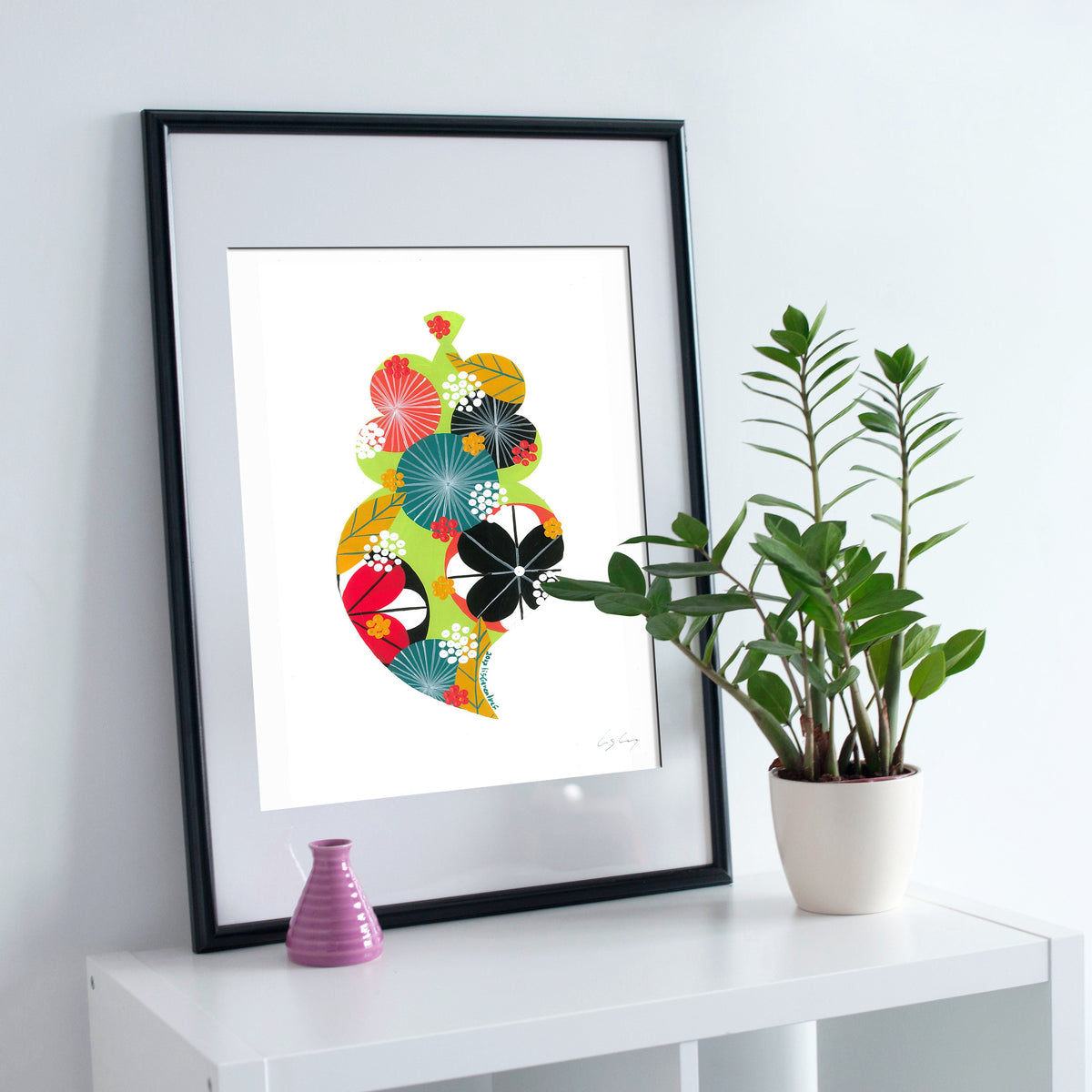 A framed fine art print, by Lis Gonçalves. Open edition, signed by the artist (printed in Hahnemühle matt fibre paper) featuring a colorful Portuguese Viana heart on a white wall with a plant and small vase.