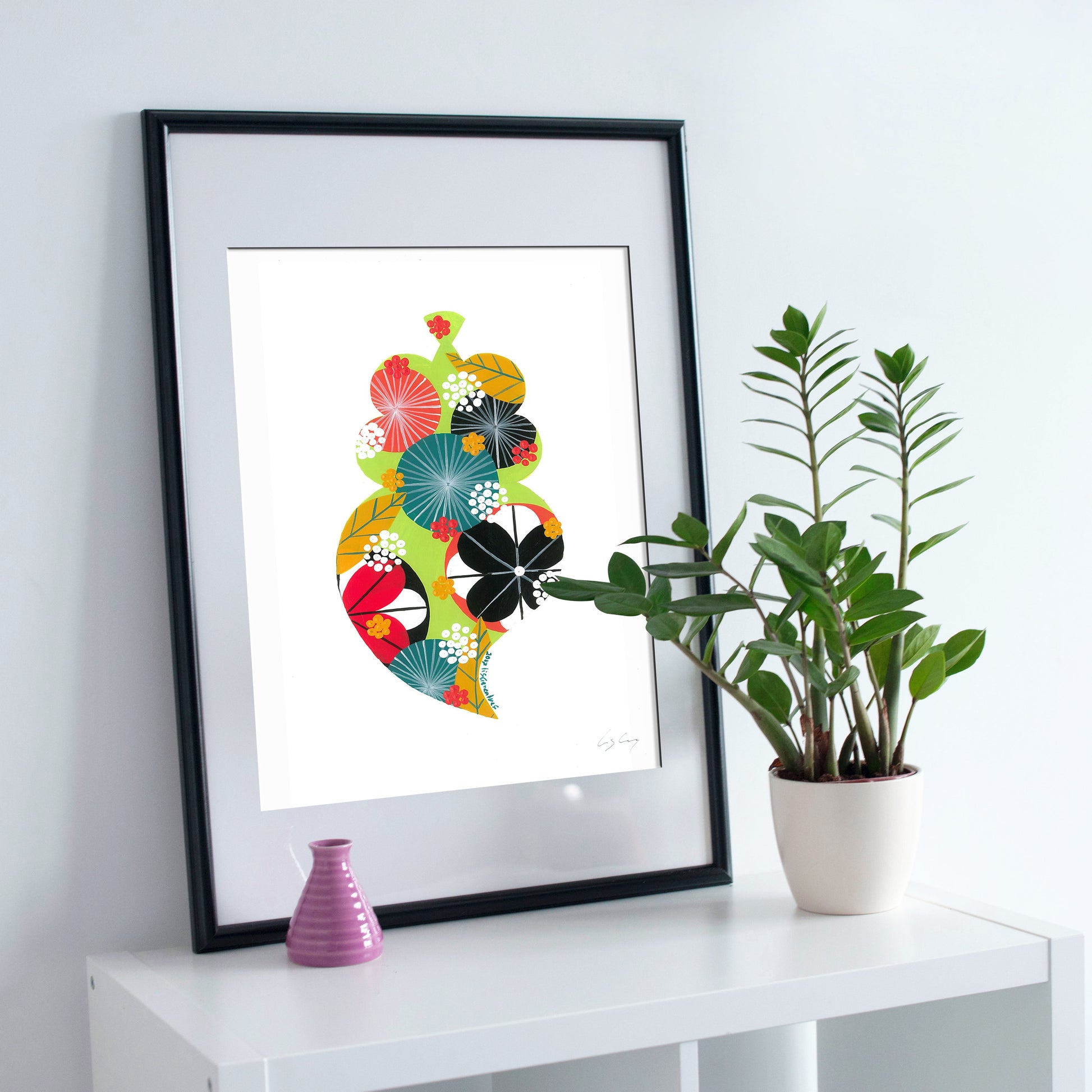 A framed fine art print, by Lis Gonçalves. Open edition, signed by the artist (printed in Hahnemühle matt fibre paper) featuring a colorful Portuguese Viana heart on a white wall with a plant and small vase.
