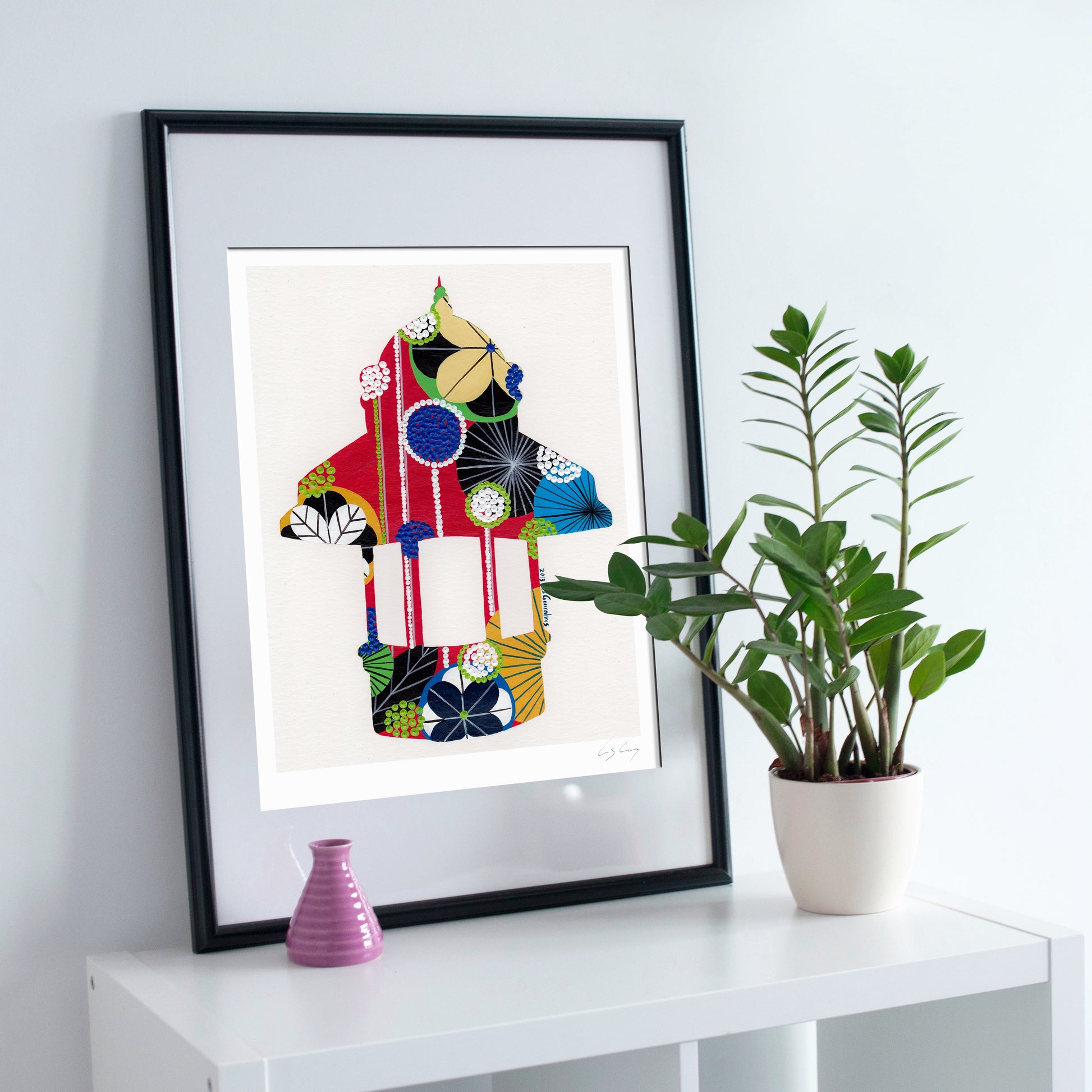 A framed fine art print, by Lis Gonçalves. Open edition, signed by the artist (printed in Hahnemühle matt fibre paper) featuring a colorful, patterned kiosk illustration leaning against a white wall on a white shelf next to a potted green plant and a small pink vase.