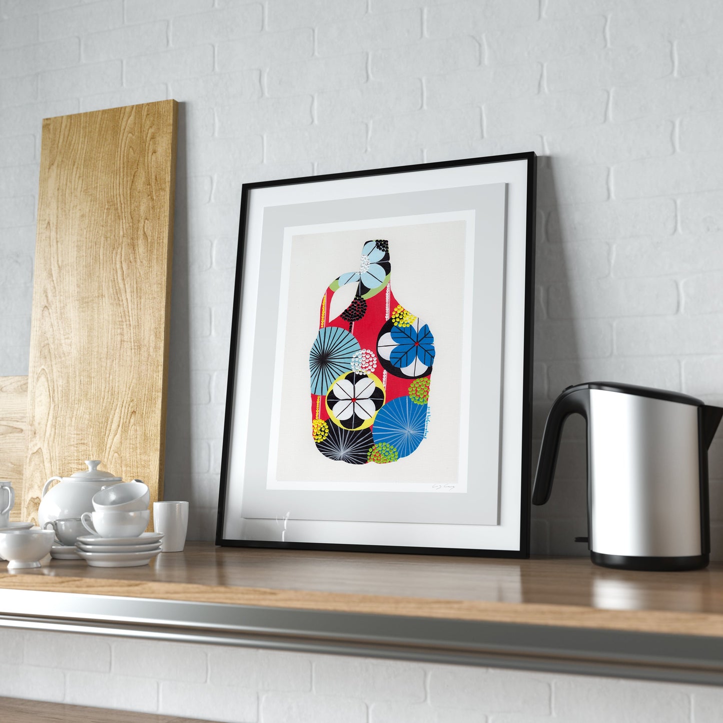 A framed fine art print, by Lis Gonçalves. Open edition, signed by the artist. Printed in Hahnemühle matt fibre paper; featuring a colorful abstract Japanese-style floral design of a wine demijohn in red, blue, yellow, black, and white, displayed on a wooden kitchen counter next to a kettle and a wooden board against a white brick wall.