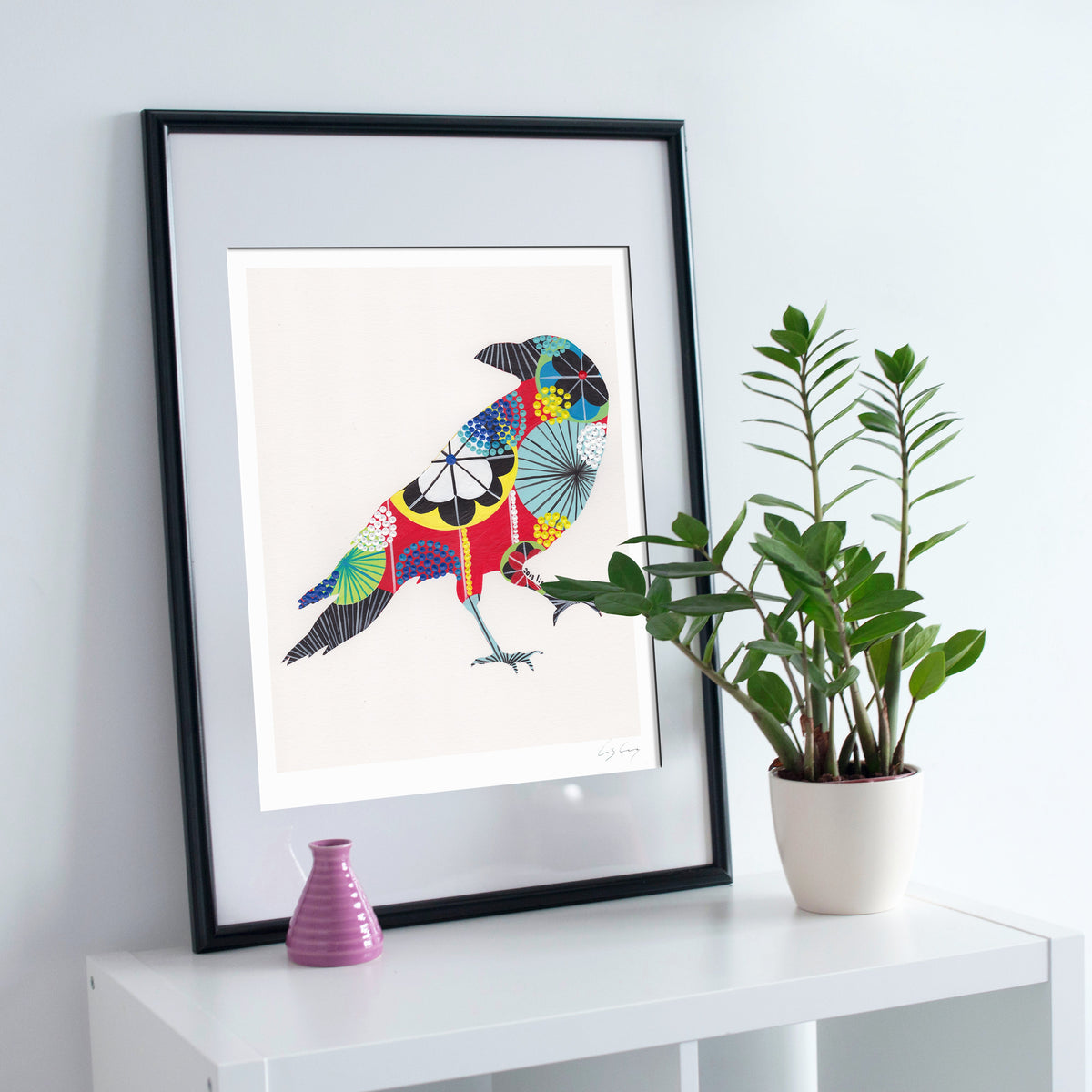 A framed fine art print, by Lis Gonçalves. Open edition, signed by the artist. Printed in Hahnemühle matt fibre paper; featuring a colorful, patterned raven on a white background, styled on a white shelf next to a potted green plant and a small pink vase against a light gray wall.