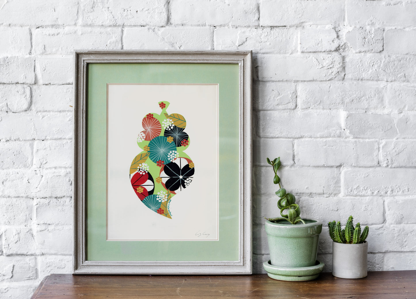 A framed fine art print, by Lis Gonçalves. Open edition, signed by the artist (printed in Hahnemühle matt fibre paper) featuring a colorful Portuguese Viana heart on a white brick wall with potted plants below.