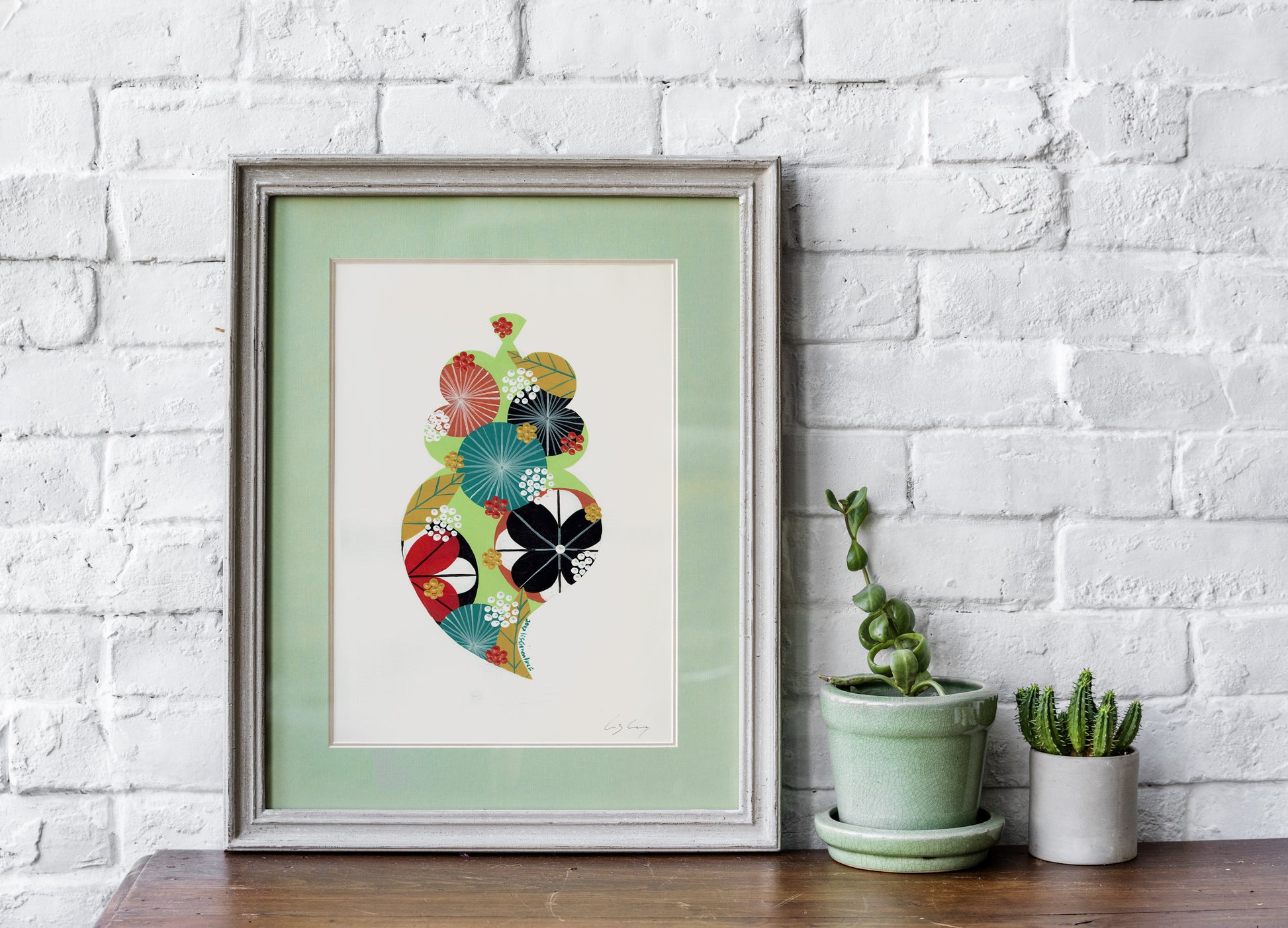 A framed fine art print, by Lis Gonçalves. Open edition, signed by the artist (printed in Hahnemühle matt fibre paper) featuring a colorful Portuguese Viana heart on a white brick wall with potted plants below.