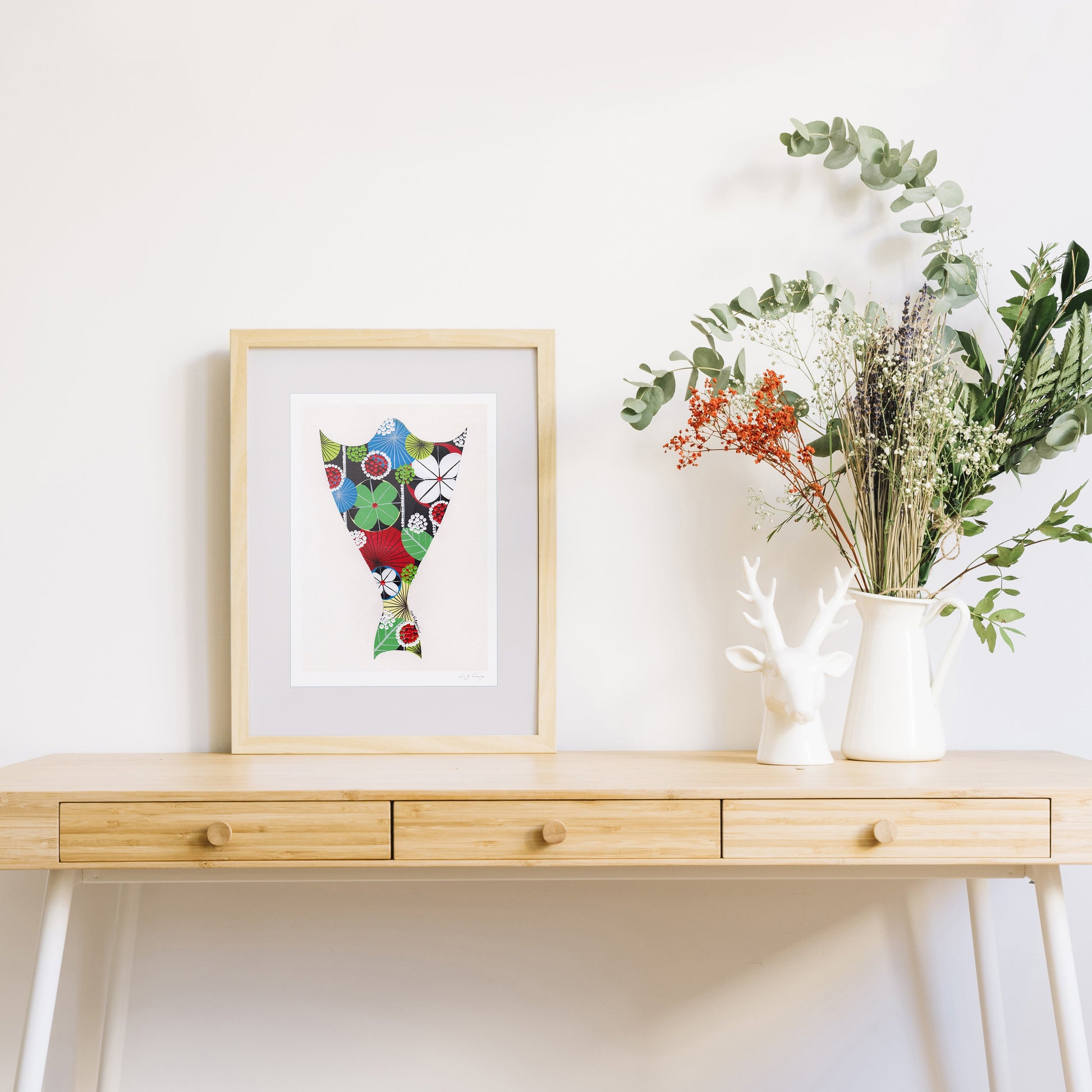 A framed fine art print, by Lis Gonçalves. Open edition, signed by the artist (printed in Hahnemühle matt fibre paper) featuring a bacalhau on a wooden desk with a vase of flowers on a white background.