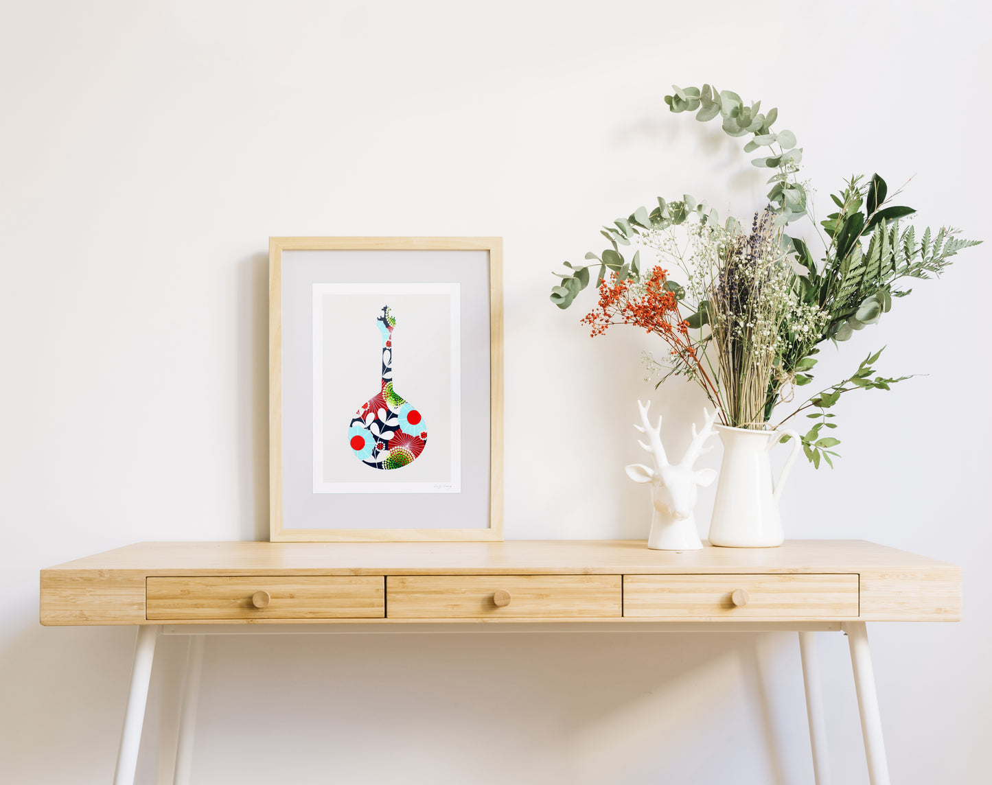 A framed fine art print, by Lis Gonçalves. Open edition, signed by the artist (printed in Hahnemühle matt fibre paper) featuring a colorful Portuguese guitar, on a wooden table with a vase of flowers on a white background.
