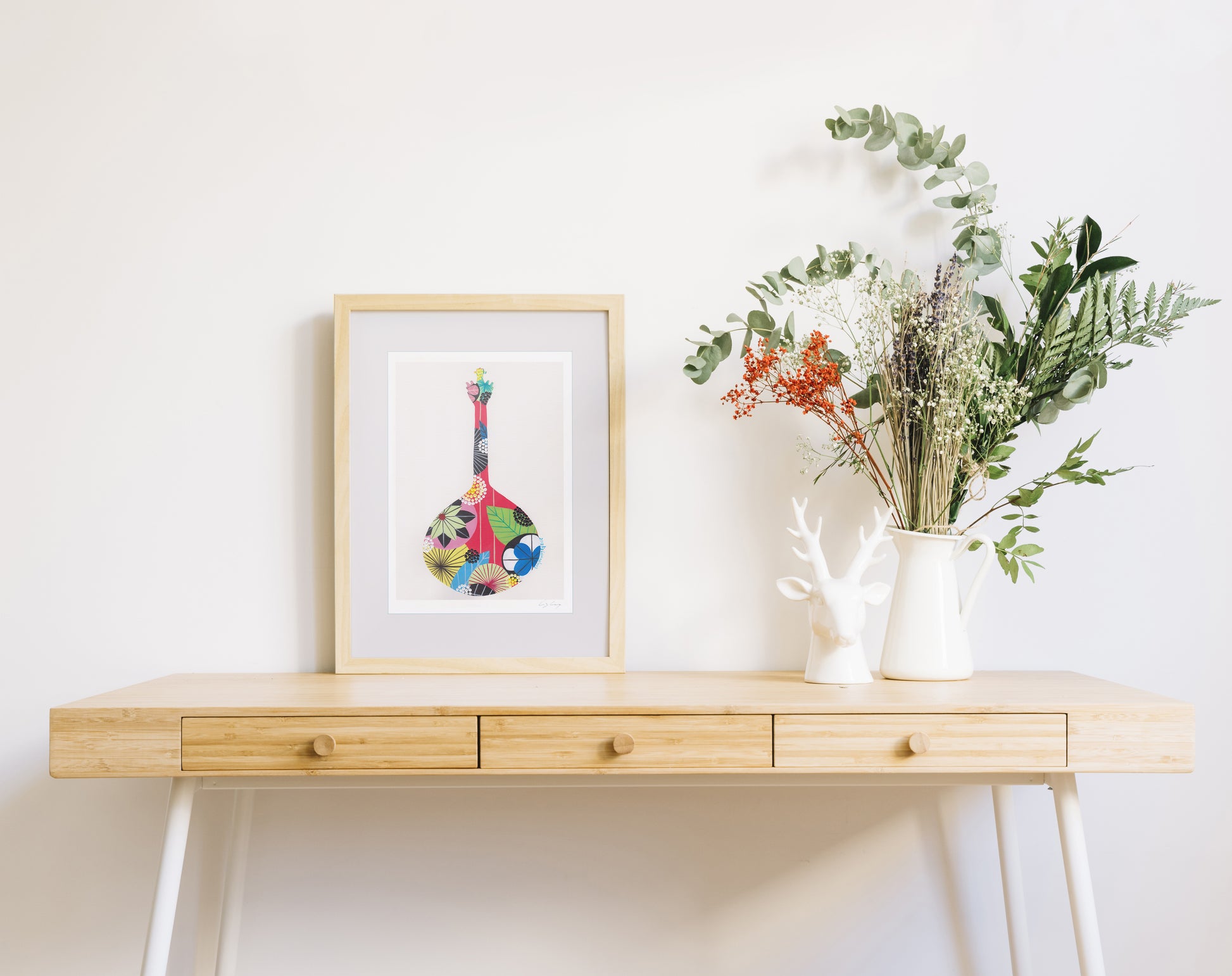 A framed fine art print, by Lis Gonçalves. Open edition, signed by the artist (printed in Hahnemühle matt fibre paper) featuring a colorful Portuguese guitar on a wooden desk with a vase of flowers on a white wall background