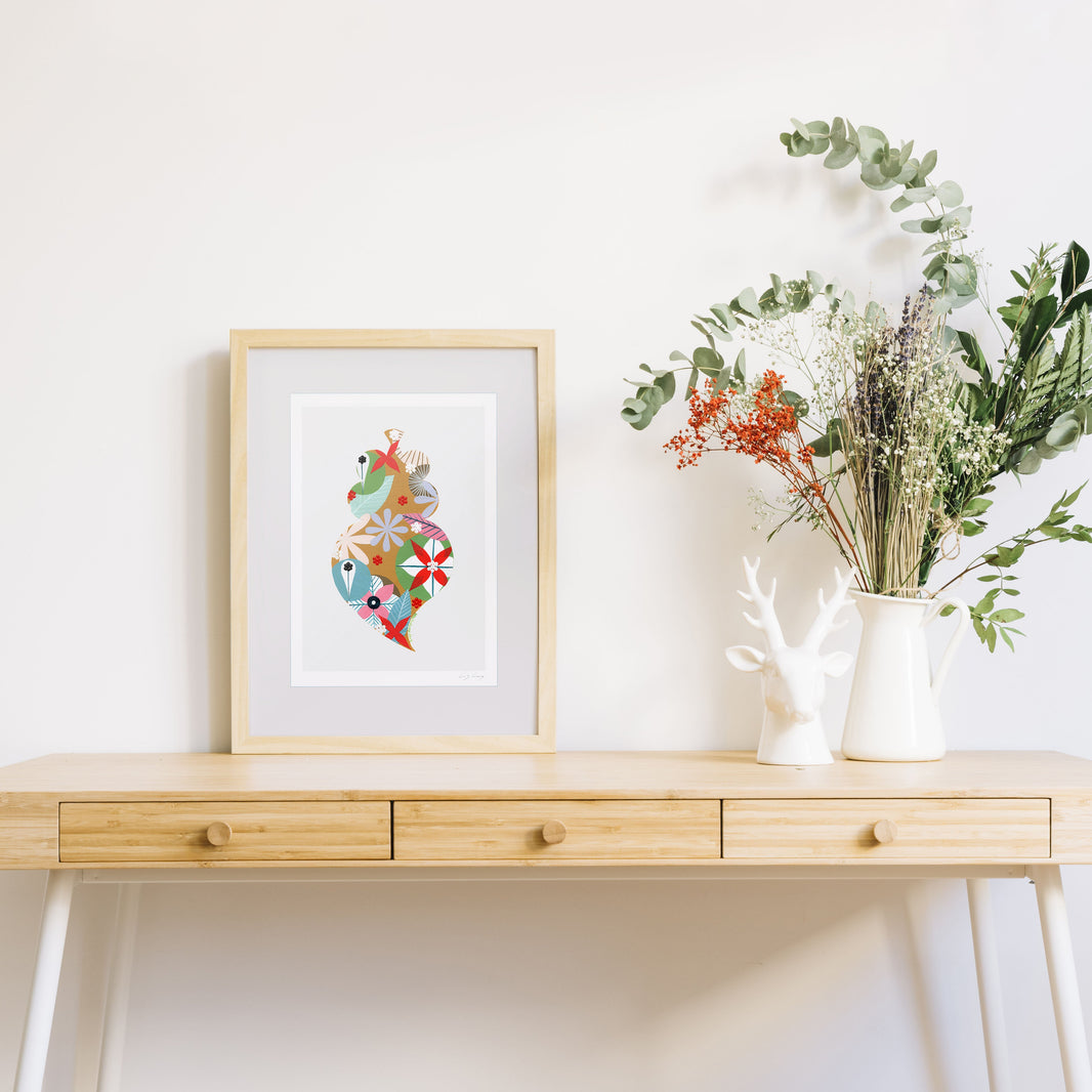 A framed fine art print, by Lis Gonçalves. Open edition, signed by the artist (printed in Hahnemühle matt fibre paper) featuring a colorful Portuguese Viana heart on a wooden desk with a framed artwork and a vase of flowers on a white background