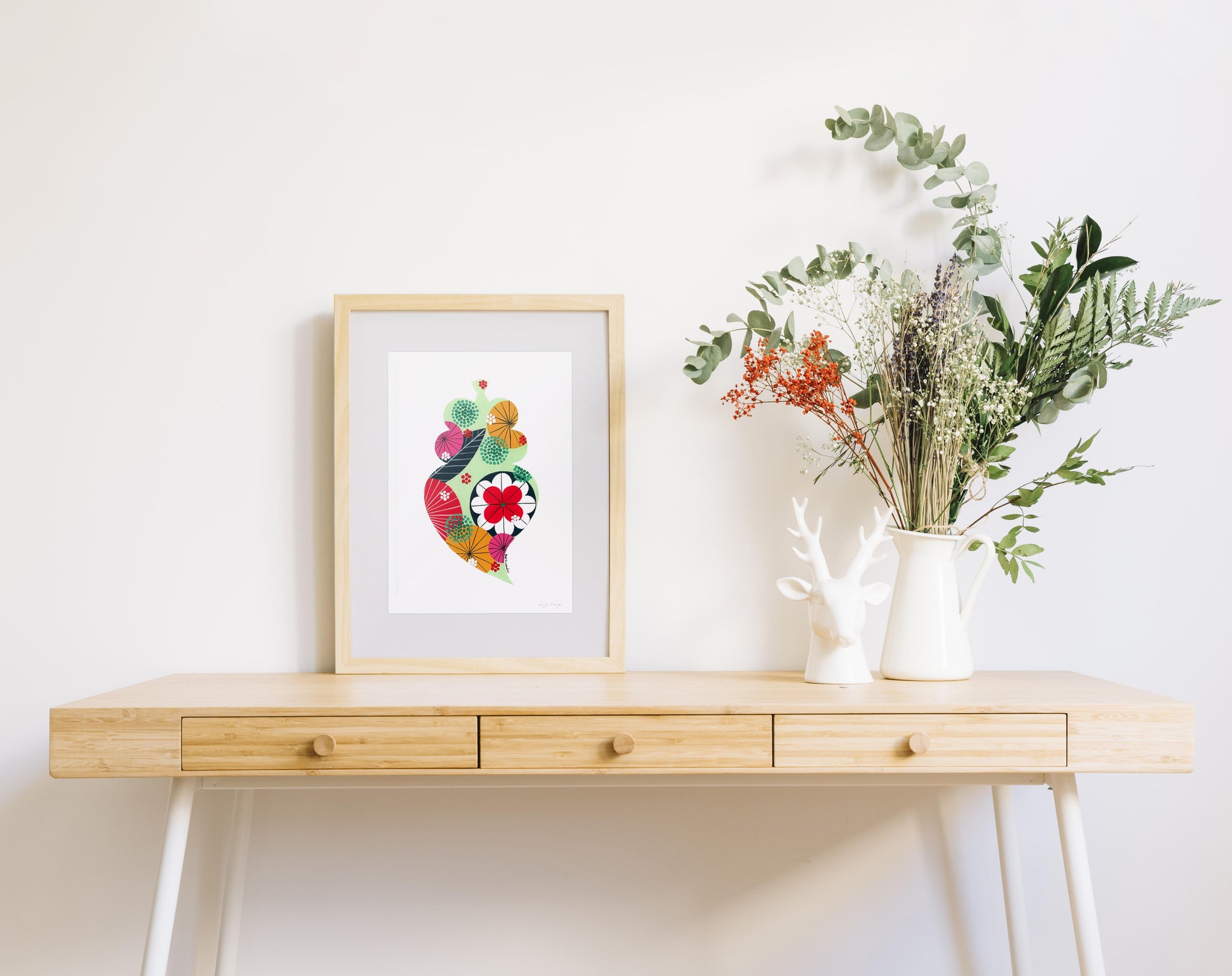 A framed fine art print, by Lis Gonçalves. Open edition, signed by the artist (printed in Hahnemühle matt fibre paper) featuring a colorful Portuguese Viana heart on a wooden table with a vase of flowers against a white wall