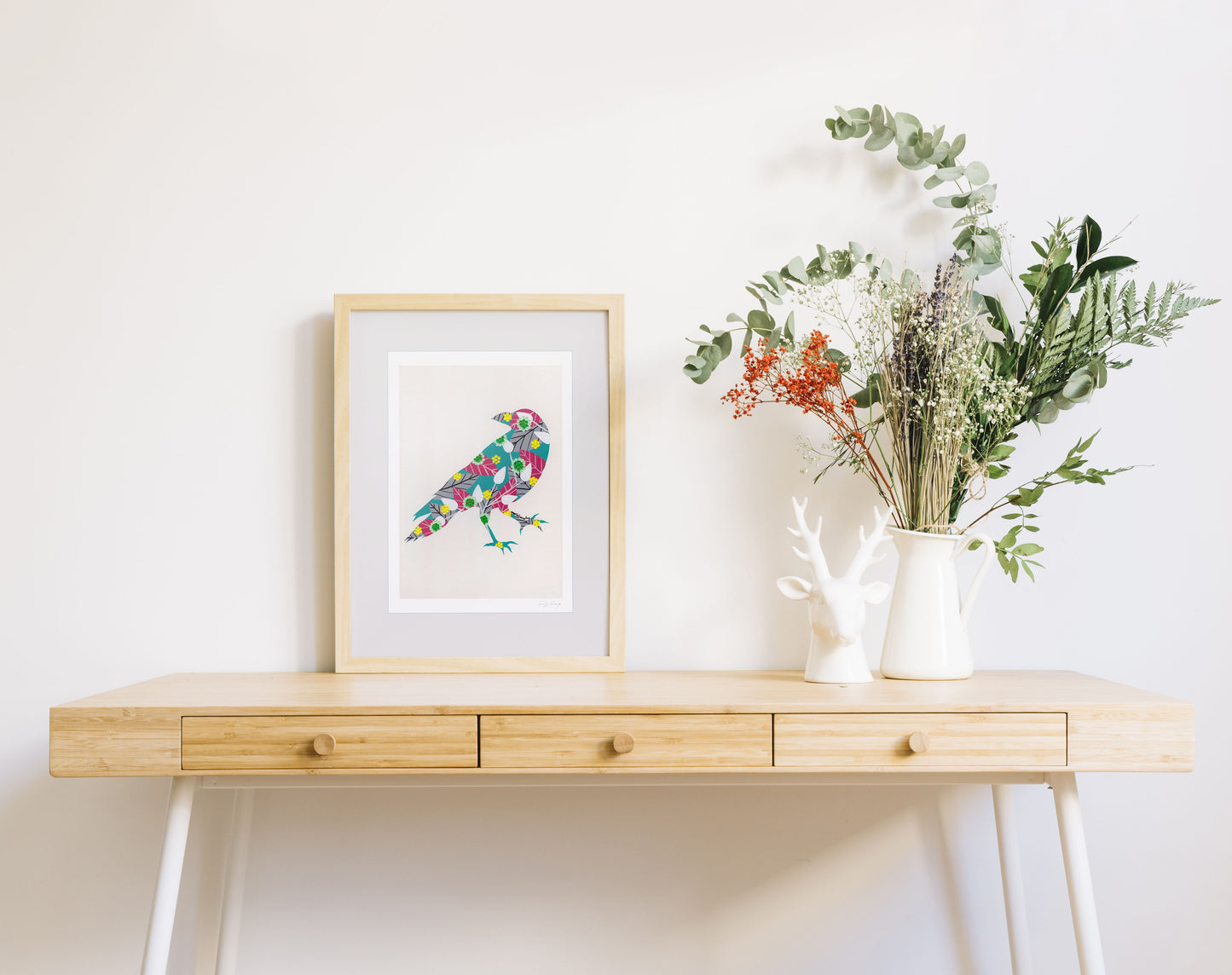 A framed fine art print, by Lis Gonçalves. Open edition, signed by the artist (printed in Hahnemühle matt fibre paper) featuring a raven on a wooden desk with a vase of flowers on a white background