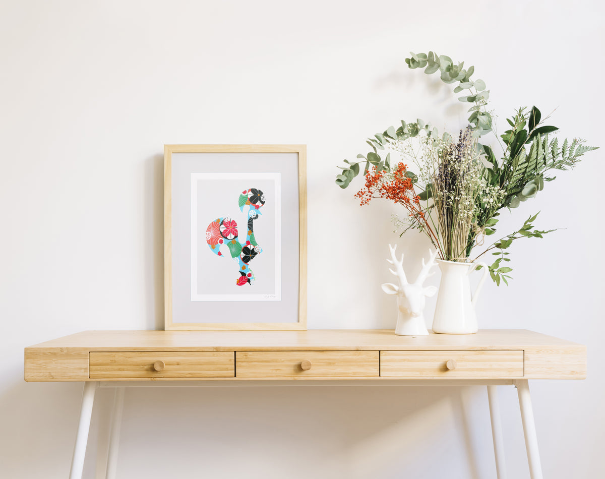 A framed fine art print, by Lis Gonçalves. Open edition, signed by the artist (printed in Hahnemühle matt fibre paper) featuring a colorful Portuguese rooster on a wooden desk with a vase of flowers on a white wall background
