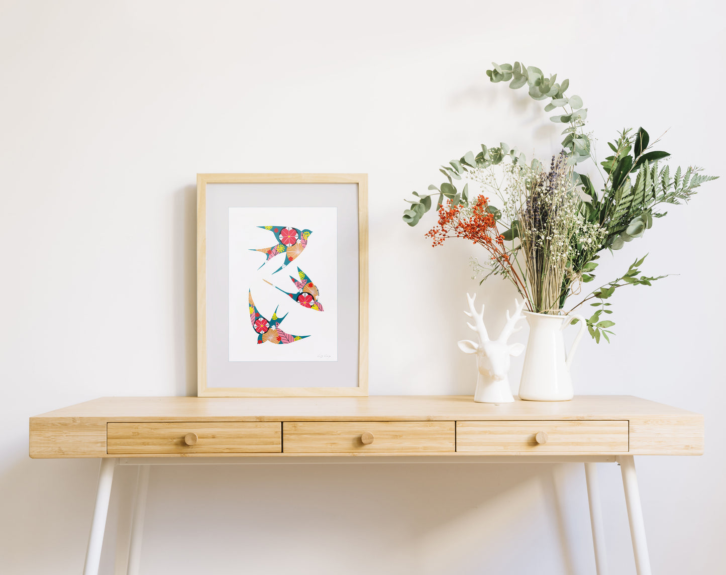 A framed fine art print, by Lis Gonçalves. Open edition, signed by the artist (printed in Hahnemühle matt fibre paper) featuring colorful swallows on a wooden desk with a vase of flowers on a white background