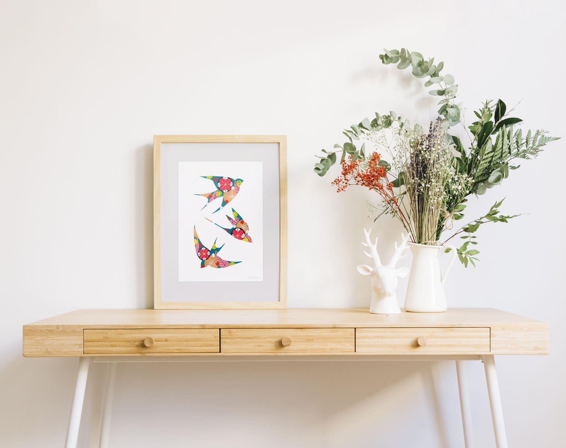 A framed fine art print, by Lis Gonçalves. Open edition, signed by the artist (printed in Hahnemühle matt fibre paper) featuring colorful swallows on a wooden desk with a vase of flowers on a white background