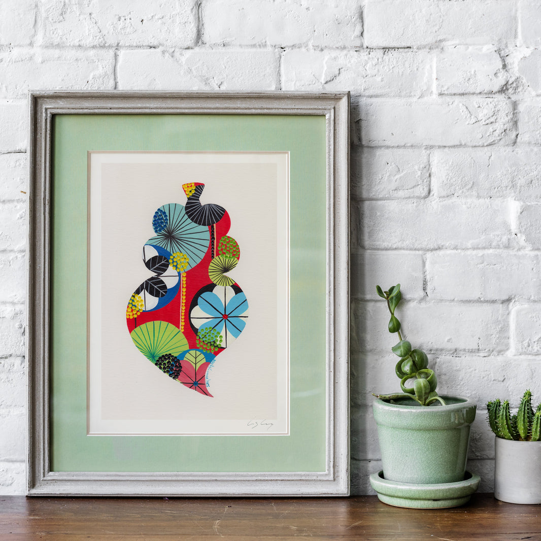 A framed fine art print, by Lis Gonçalves. Open edition, signed by the artist (printed in Hahnemühle matt fibre paper) featuring a colorful Portuguese "Viana" heart on a white background, styled on a wooden shelf against a white brick wall with two small potted plants.