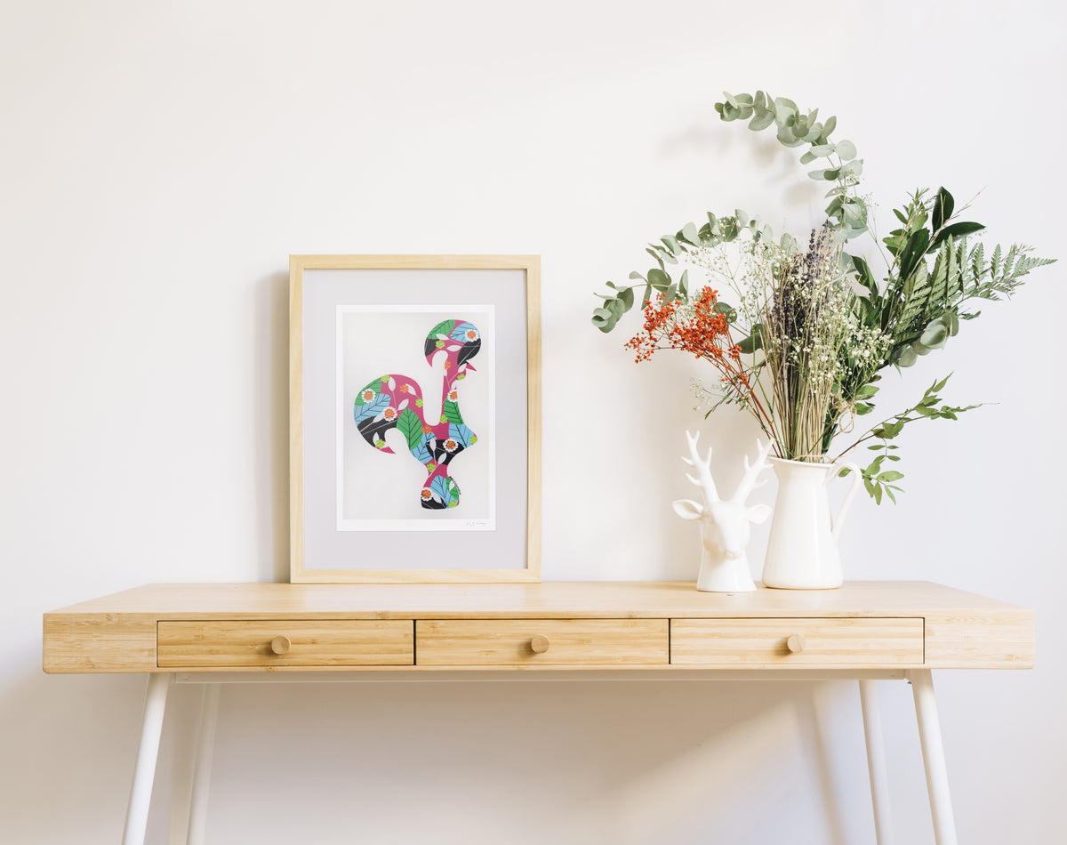 A framed fine art print, by Lis Gonçalves. Open edition, signed by the artist (printed in Hahnemühle matt fibre paper) featuring a colorful Portuguese rooster on a wooden desk with a vase of flowers on a white background