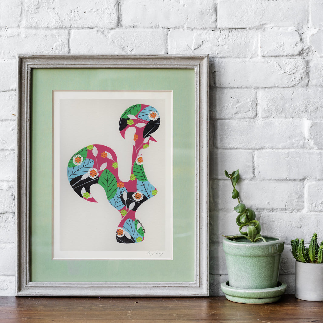 A framed fine art print, by Lis Gonçalves. Open edition, signed by the artist (printed in Hahnemühle matt fibre paper) featuring a colorful Portuguese rooster with floral and leaf patterns, displayed on a wooden surface against a white brick wall next to small potted plants.