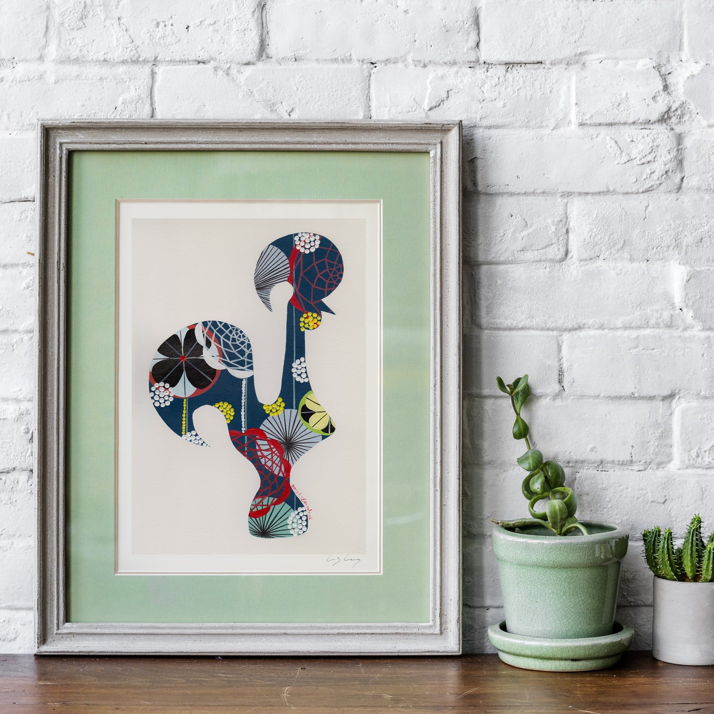 A framed fine art print, by Lis Gonçalves. Open edition, signed by the artist (printed in Hahnemühle matt fibre paper) featuring a colorful Portuguese rooster, leaning against a white brick wall on a wooden surface next to two potted green plants.