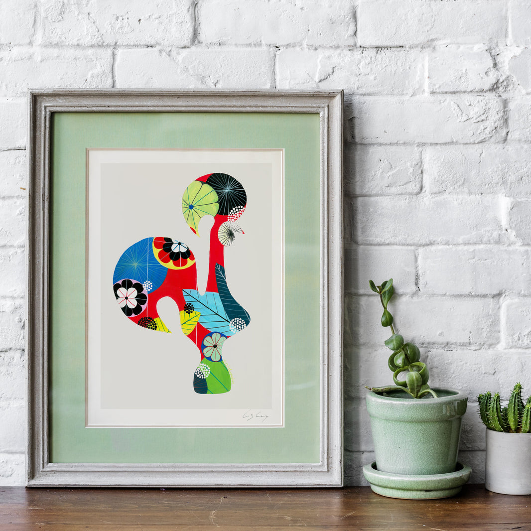 A framed fine art print, by Lis Gonçalves. Open edition, signed by the artist (printed in Hahnemühle matt fibre paper) featuring a colorful Portuguese rooster, styled on a wooden shelf against a white brick wall next to two small potted plants.