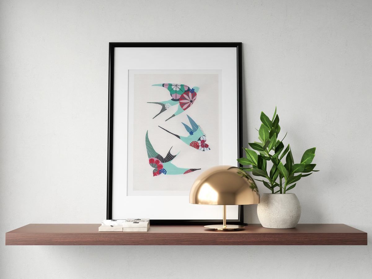 A framed fine art print, by Lis Gonçalves. Open edition, signed by the artist (printed in Hahnemühle matt fibre paper) featuring colorful swallows on a wooden shelf with a lamp and plant against a white wall