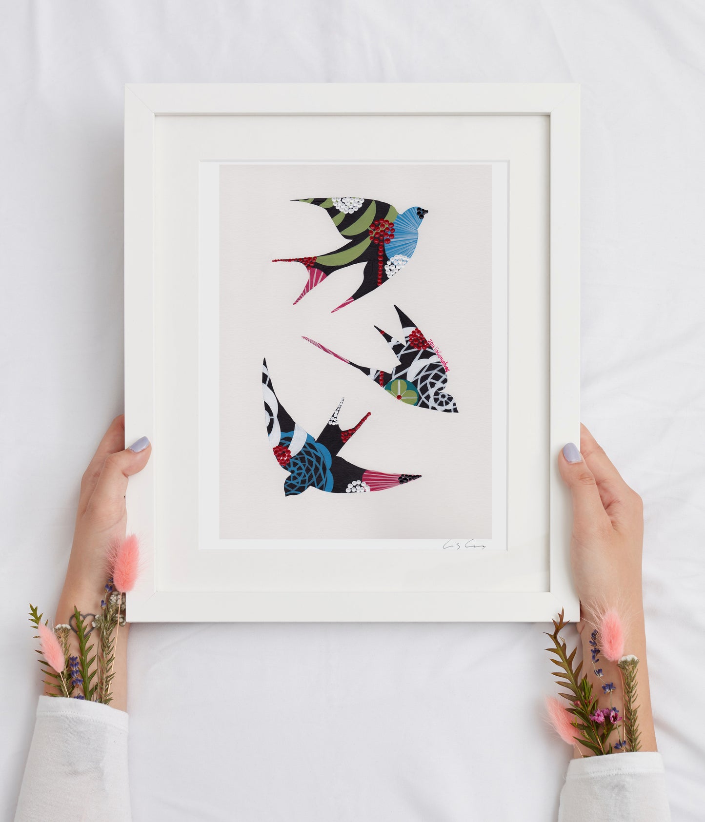 A framed fine art print, by Lis Gonçalves. Open edition, signed by the artist (printed in Hahnemühle matt fibre paper) featuring colorful swallows held by hands on a white background