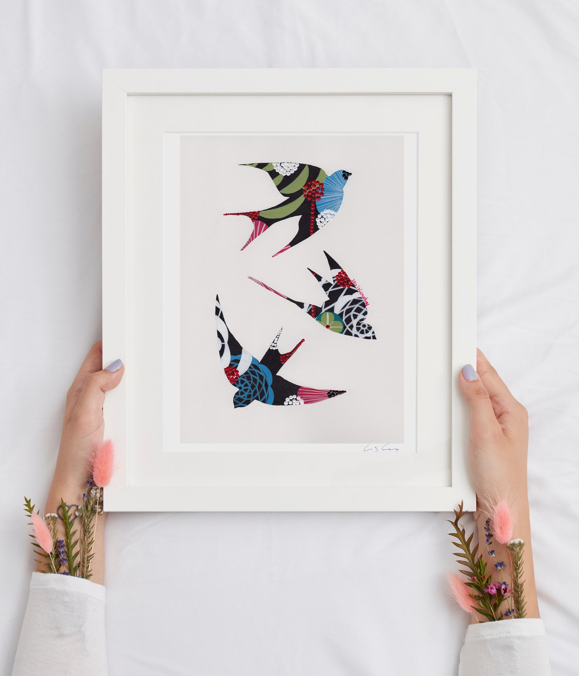 A framed fine art print, by Lis Gonçalves. Open edition, signed by the artist (printed in Hahnemühle matt fibre paper) featuring colorful swallows held by hands on a white background