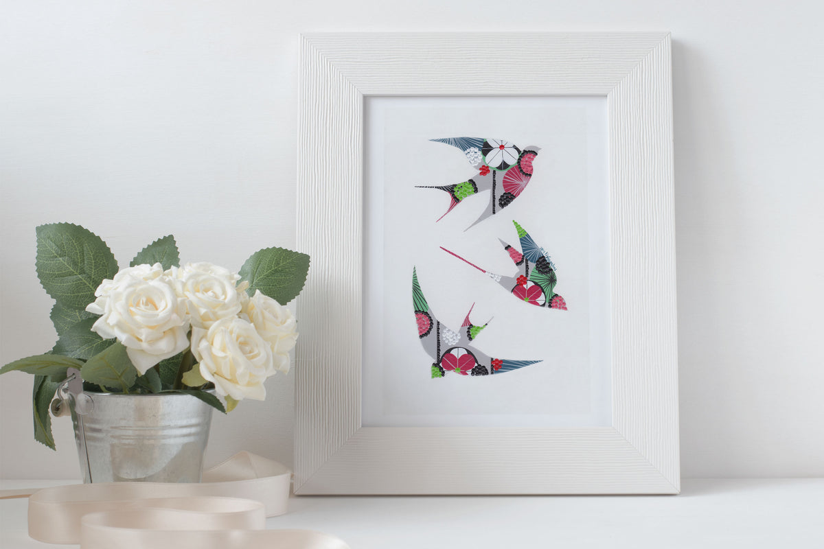 A framed fine art print, by Lis Gonçalves. Open edition, signed by the artist (printed in Hahnemühle matt fibre paper) featuring colorful swallows on a white surface with a vase of white roses.