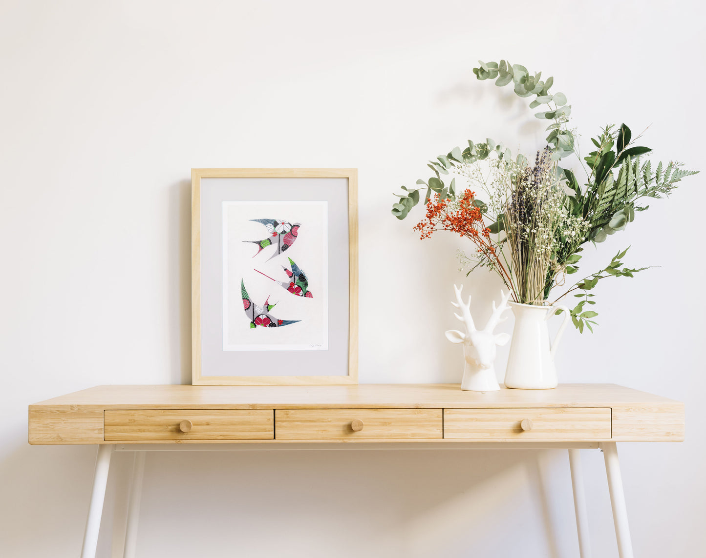 A framed fine art print, by Lis Gonçalves. Open edition, signed by the artist (printed in Hahnemühle matt fibre paper) featuring colorful swallows on a wooden desk with a vase of flowers on a white background