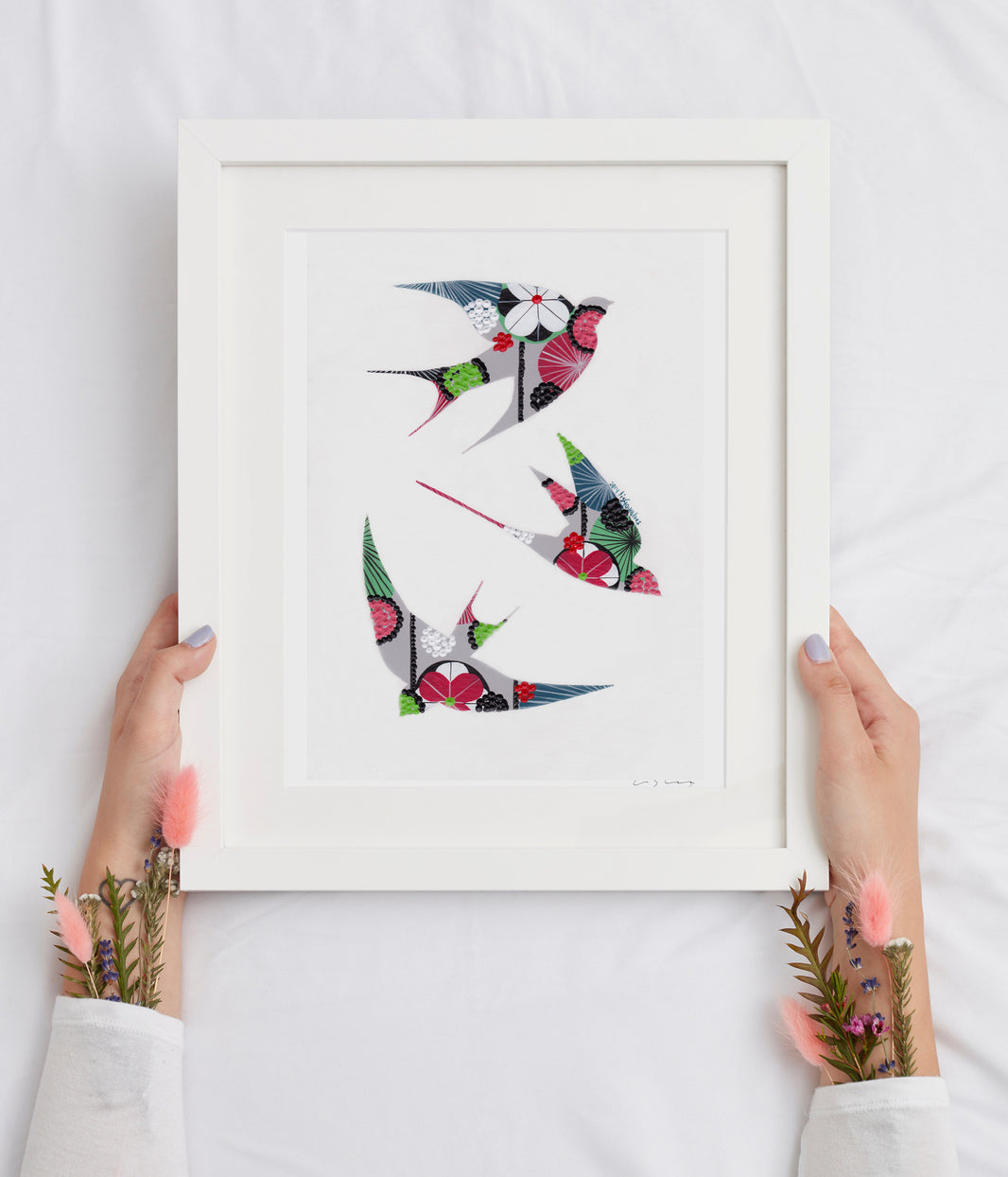 A framed fine art print, by Lis Gonçalves. Open edition, signed by the artist (printed in Hahnemühle matt fibre paper) featuring colorful swallows held by hands on a white background