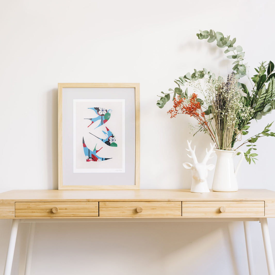 A framed fine art print, by Lis Gonçalves. Open edition, signed by the artist (printed in Hahnemühle matt fibre paper) featuring colorful swallows on a wooden desk with a vase of flowers on a white background