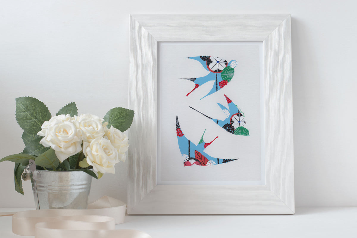 A framed fine art print, by Lis Gonçalves. Open edition, signed by the artist (printed in Hahnemühle matt fibre paper) featuring colorful swallows on a white surface with a plant.