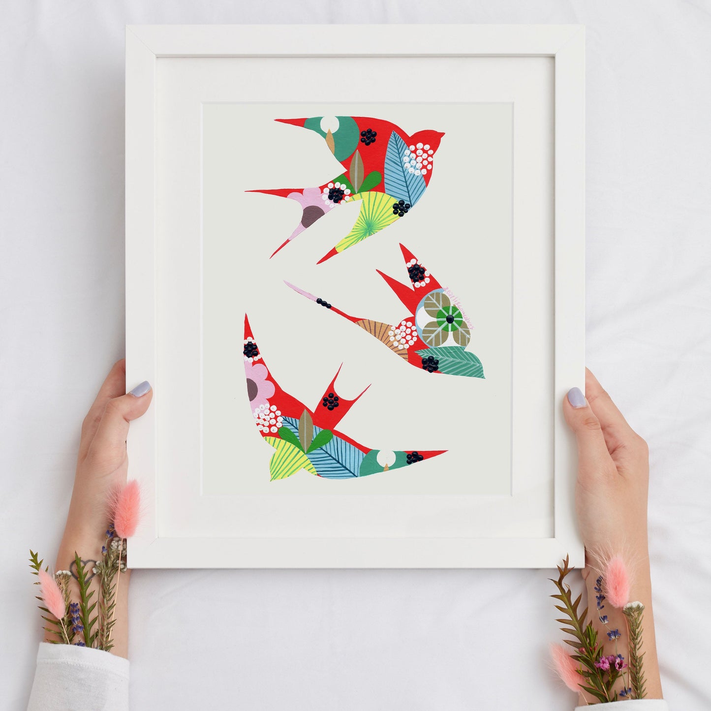 An original artwork, by Lis Gonçalves. Painted in acrylic on paper featuring colorful swallows with leaf and floral patterns, held by two hands against a white fabric background.