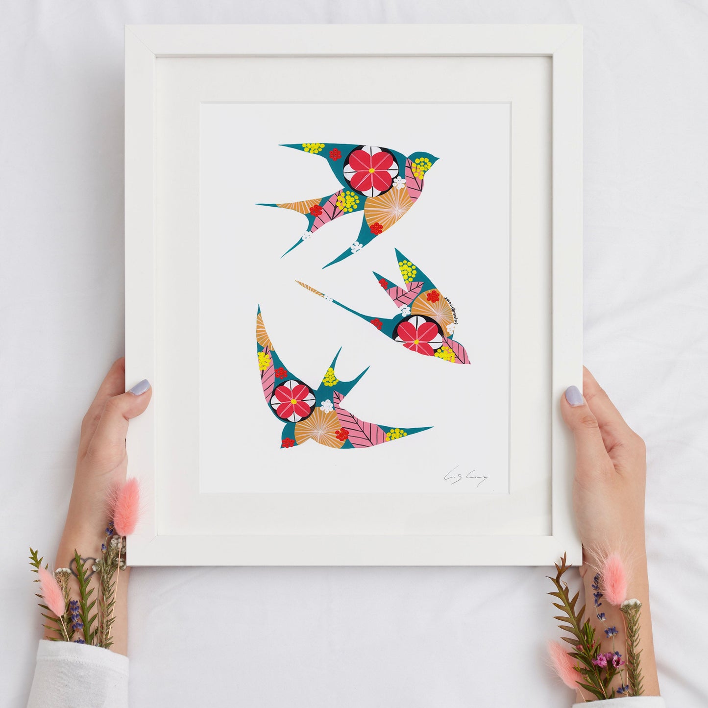 A framed fine art print, by Lis Gonçalves. Open edition, signed by the artist (printed in Hahnemühle matt fibre paper) featuring colorful swallows with floral patterns held by hands on a white background