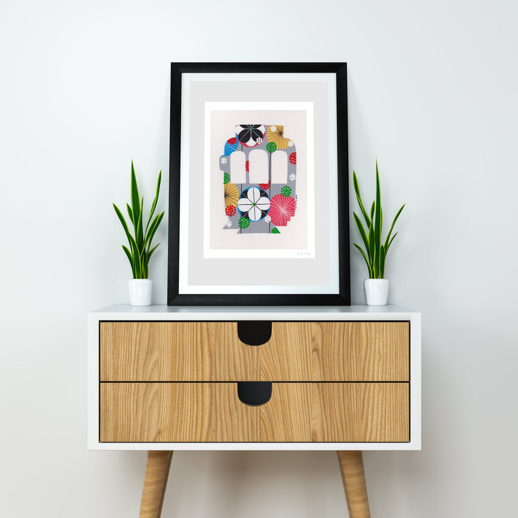 A framed fine art print, by Lis Gonçalves. Open edition, signed by the artist (printed in Hahnemühle matt fibre paper) featuring a colorful Lisbon tram, displayed on a white sideboard with two small potted green plants on either side. The print has a white mat and a black frame.