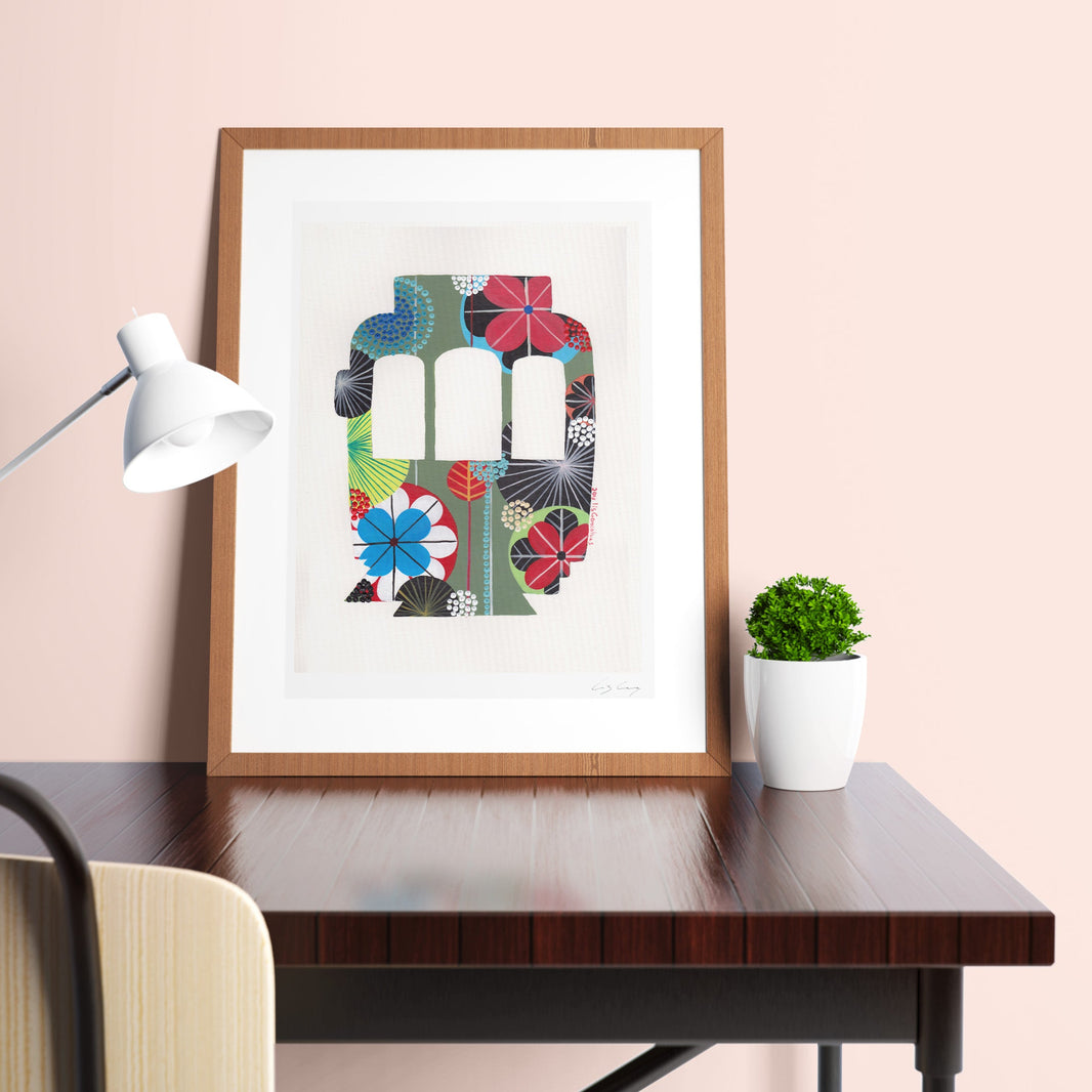 A framed fine art print, by Lis Gonçalves. Open edition, signed by the artist (printed in Hahnemühle matt fibre paper) featuring a colorful Lisbon tram on a desk with a lamp and plant.
